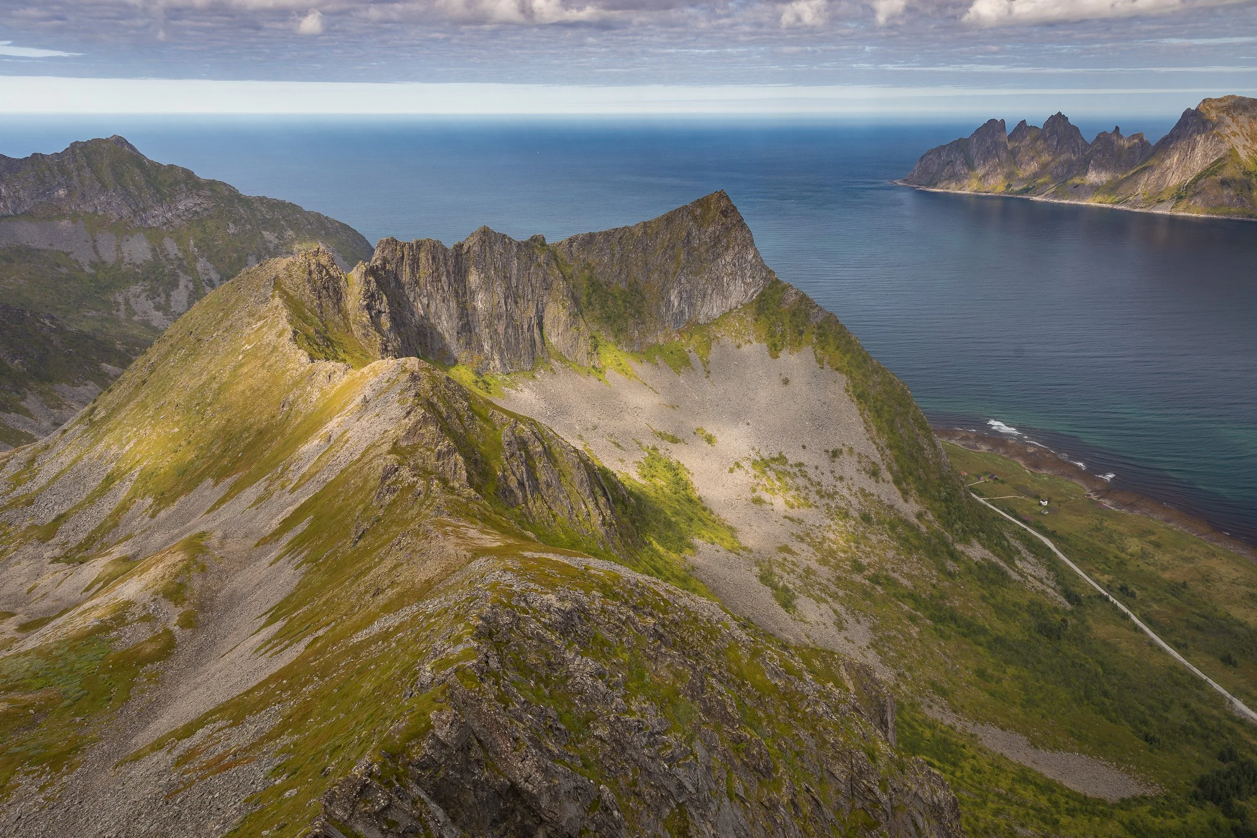 The ridge below Husfjellet runs towards the Norwegian Sea, with distant peaks and a narrow coastal road tracing the edge of the landscape.