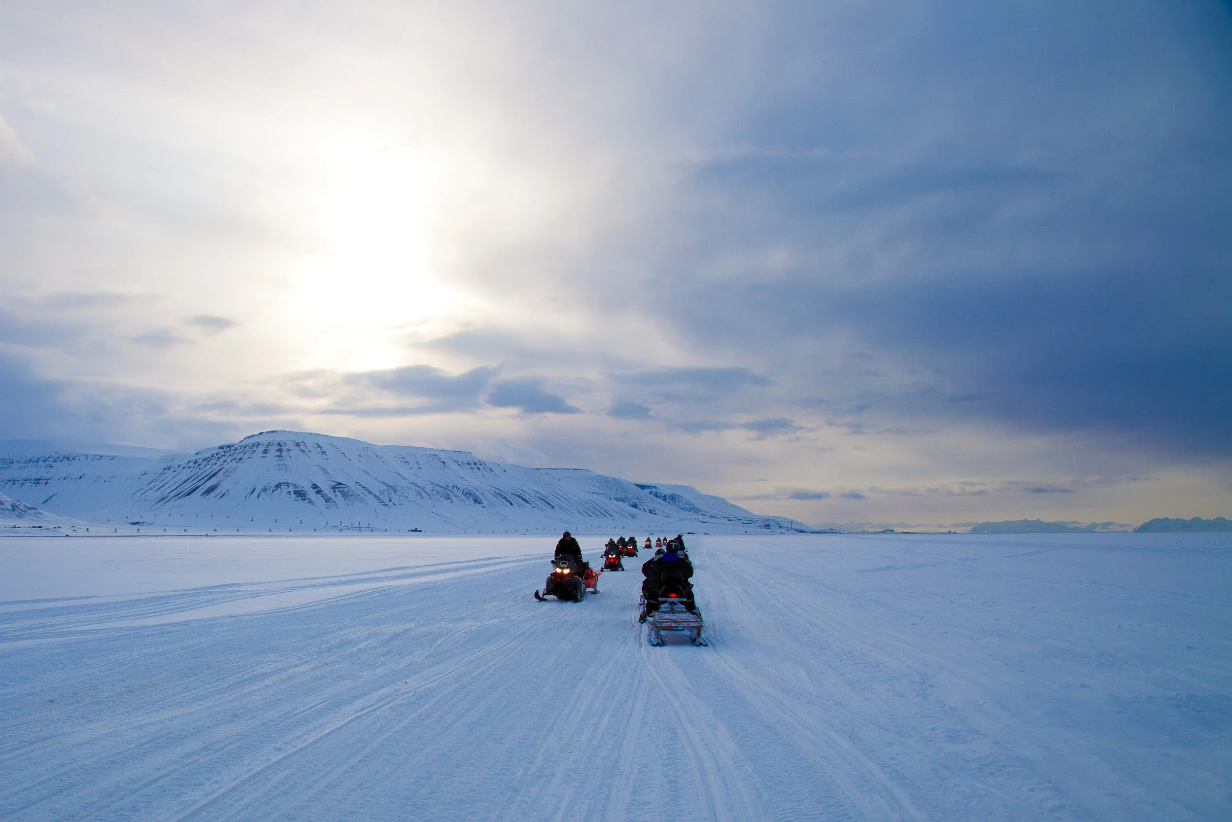 A line of snowmobiles crosses Adventdalen under cold light and a vast Svalbard sky.