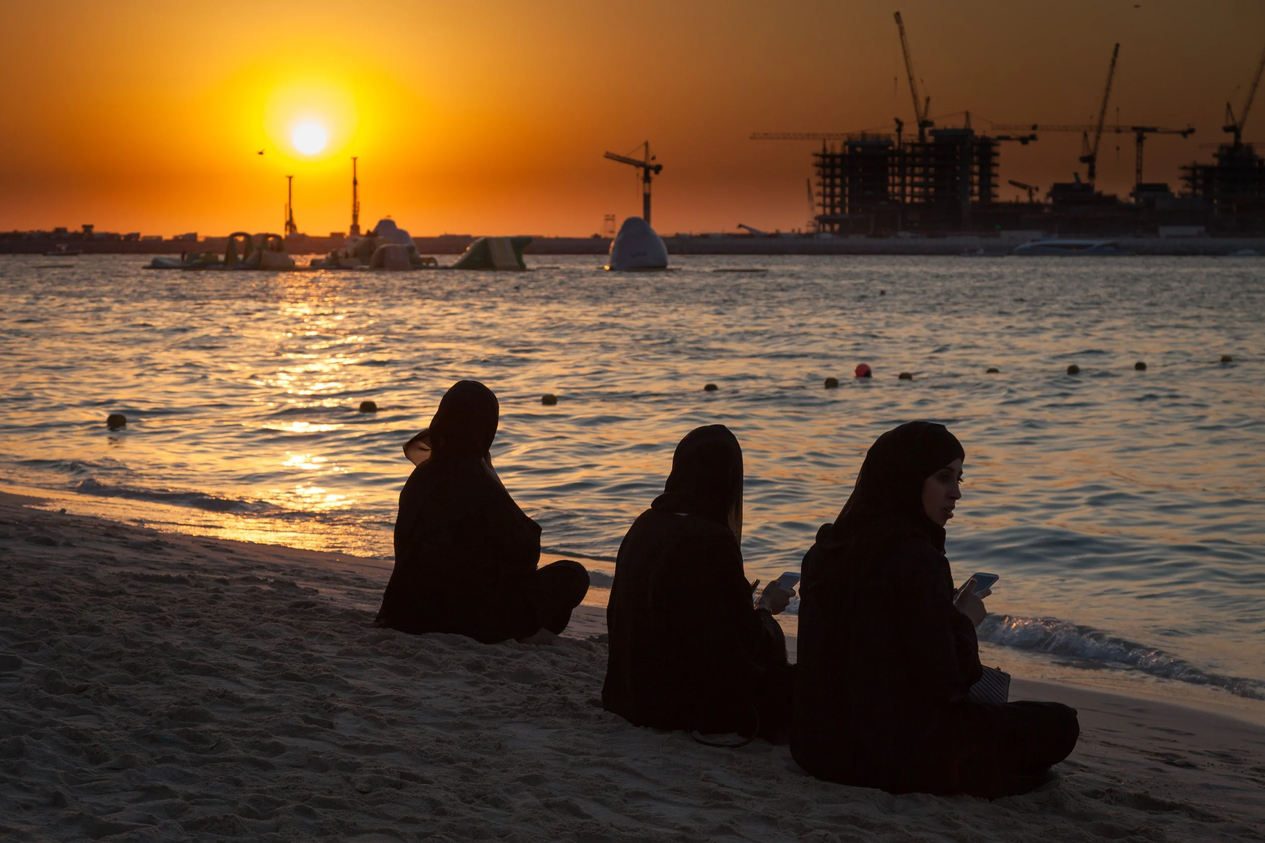 Silhouetted figures on a Dubai beach at sunset, with glowing water and cranes on the horizon.