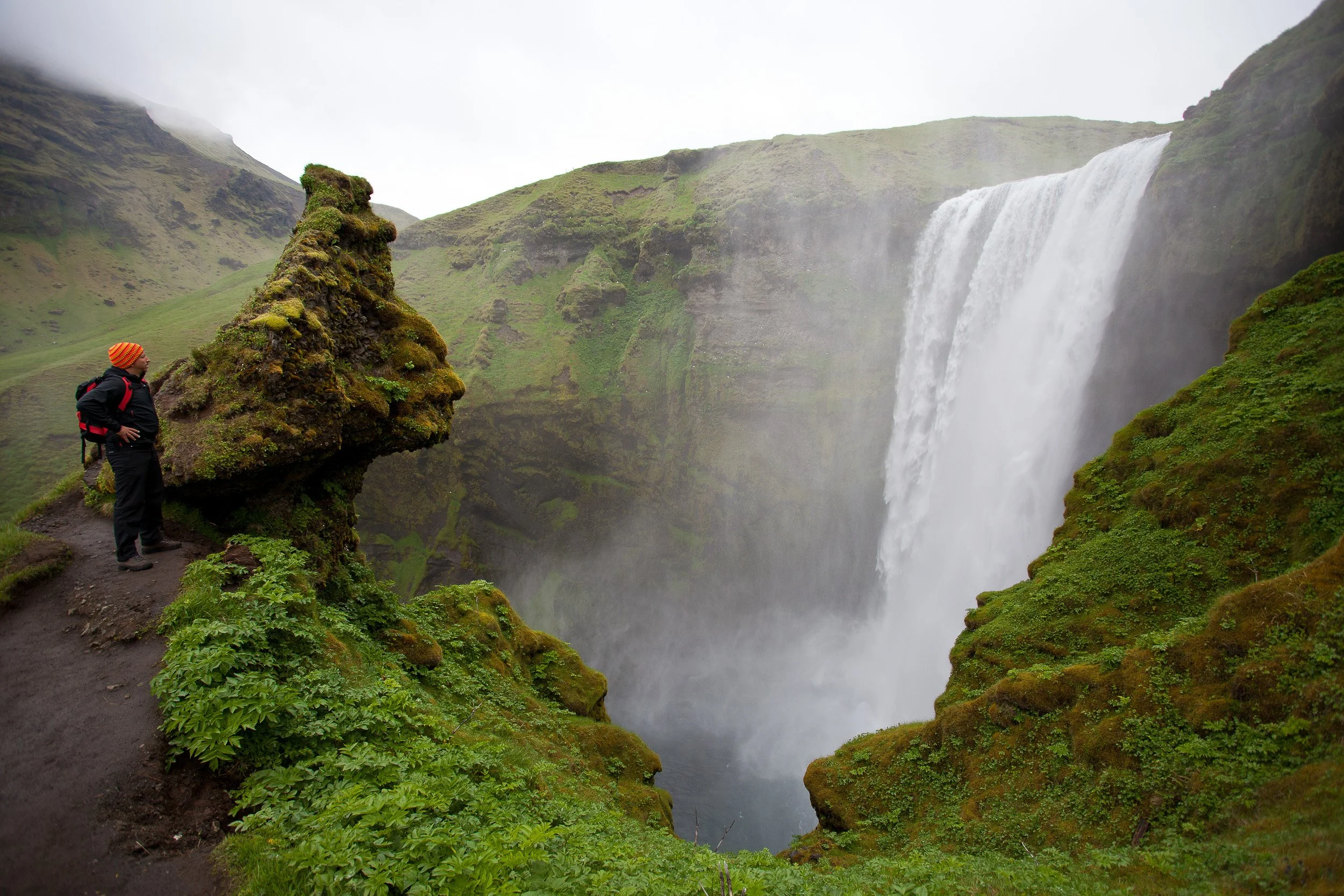 A lone hiker pauses beside the mossy trail as Skógafoss crashes into the gorge, filling the air with cold mist and the steady roar of water.