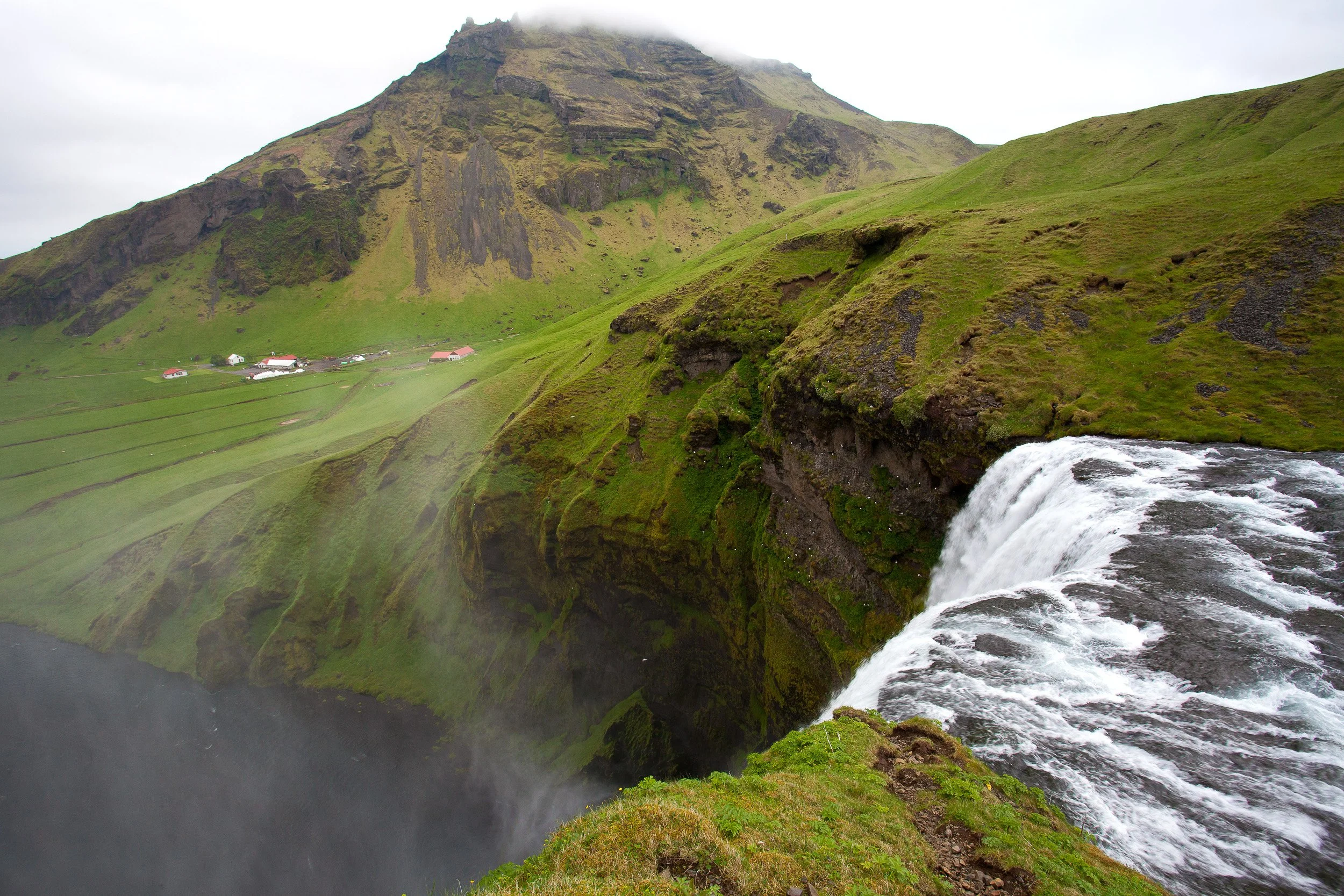 From the brink above Skógafoss, the river gathers speed before plunging into open farmland, with the valley and distant mountains fading into low cloud.