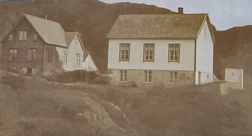 Historisk fotografi av det hvite skolehuset i Husevåg til høyre og det mørke trehuset til venstre, der Ivar Husevåg vokste opp.