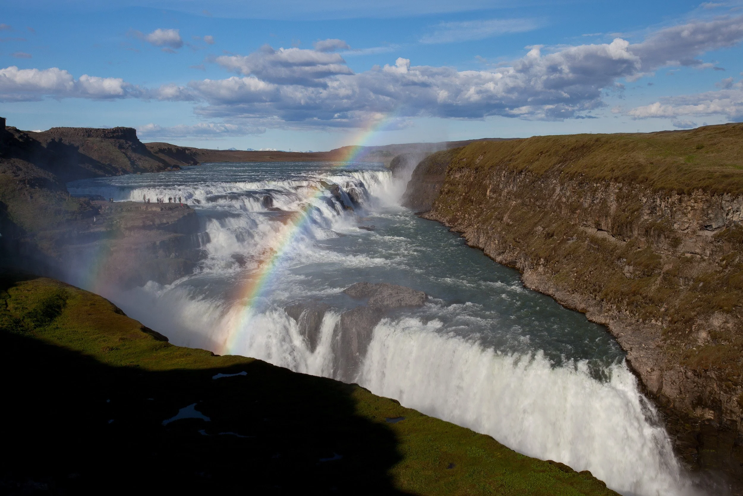 Gullfoss thunders through its rocky canyon in two great steps, sunlight catching the spray to paint a bright rainbow across the gorge.