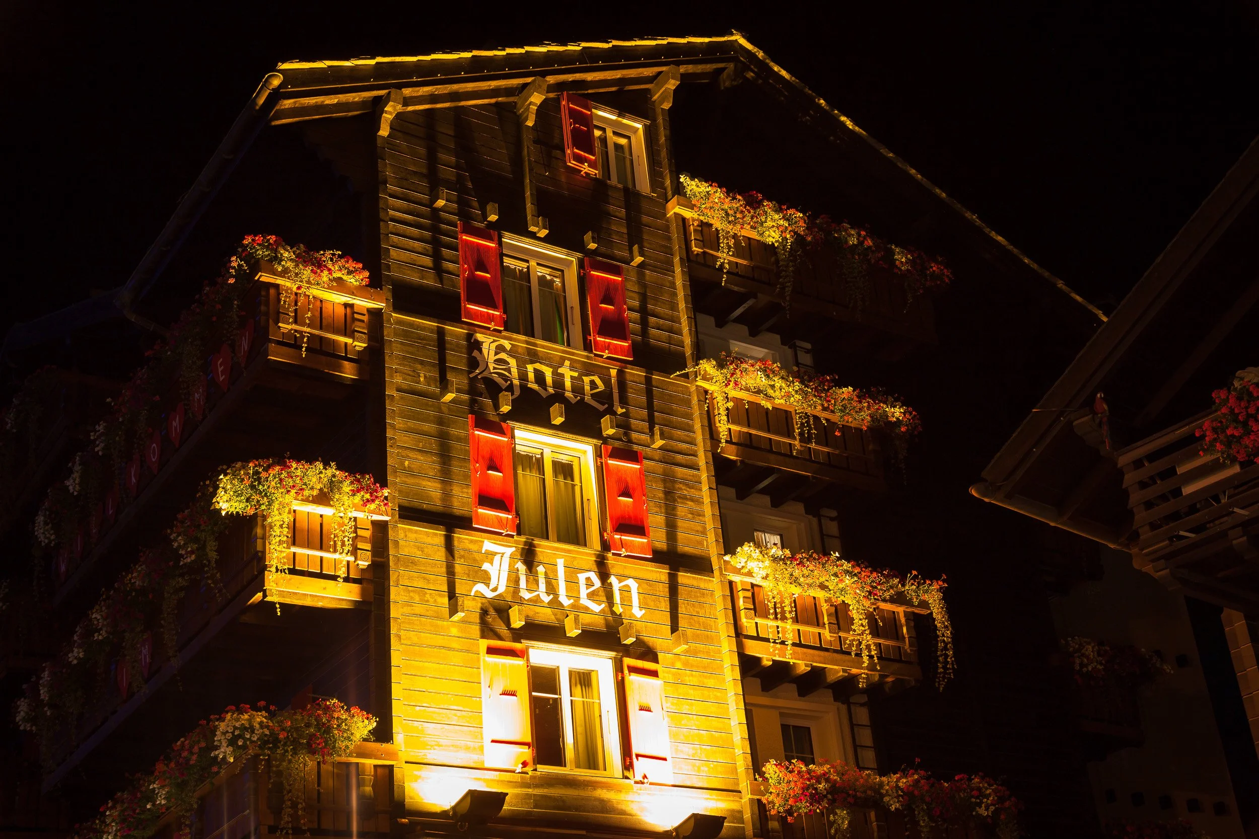 Cosy wooden façade of Hotel Julen in Zermatt glowing warmly at night, with red shutters and flower-filled balconies.