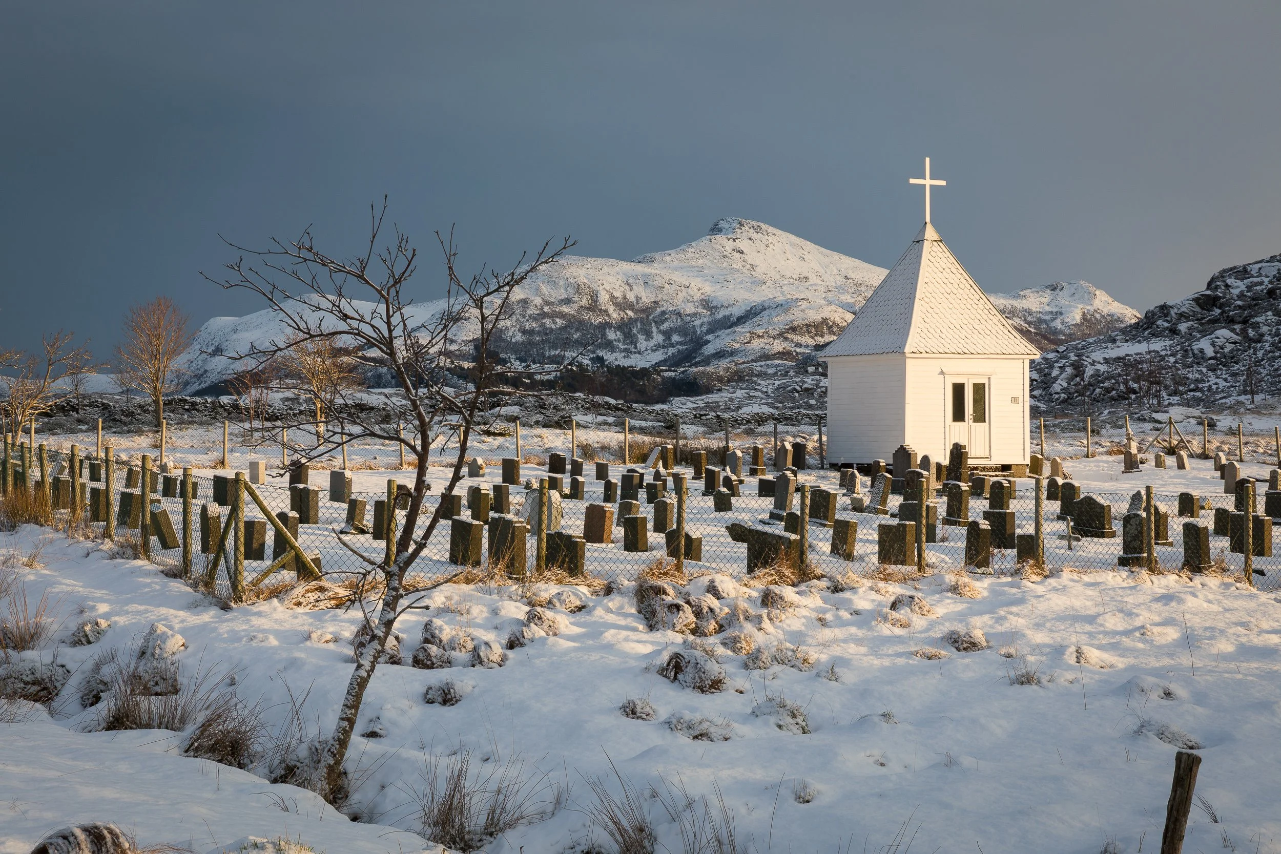 Gravplassen i Tytingvåg vinterstid, med klokkehuset, gravstøtter og snødekte fjell i bakgrunnen.