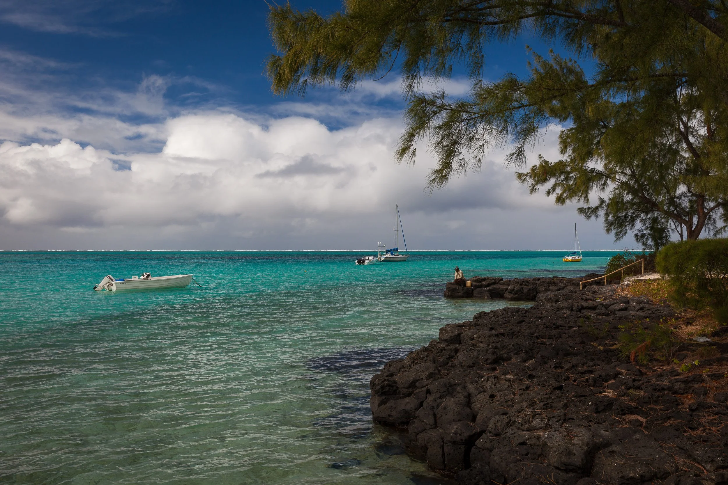 Local fisherman sitting on black lava rocks at Pointe d’Esny, Mauritius, looking out over the calm turquoise lagoon and anchored boats.