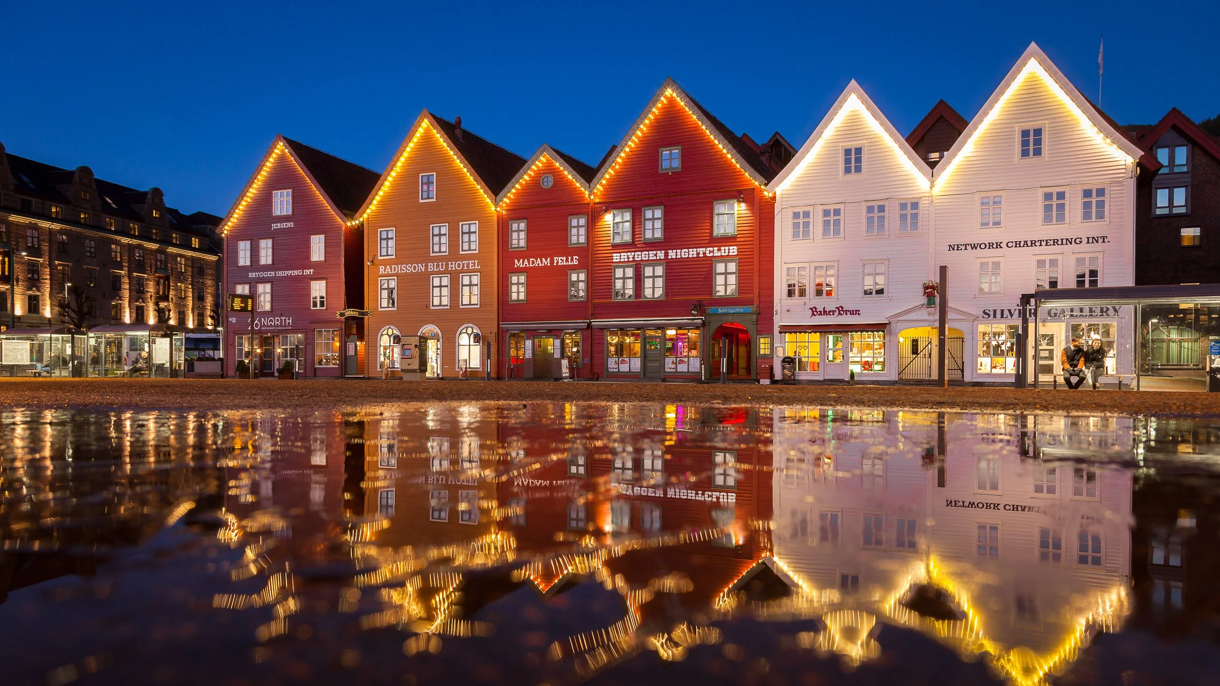 My hometown Bergen in Western Norway – the colourful wooden houses of Bryggen glowing in blue-hour light, their festive lights and façades reflected in a rain puddle on the cobblestones.