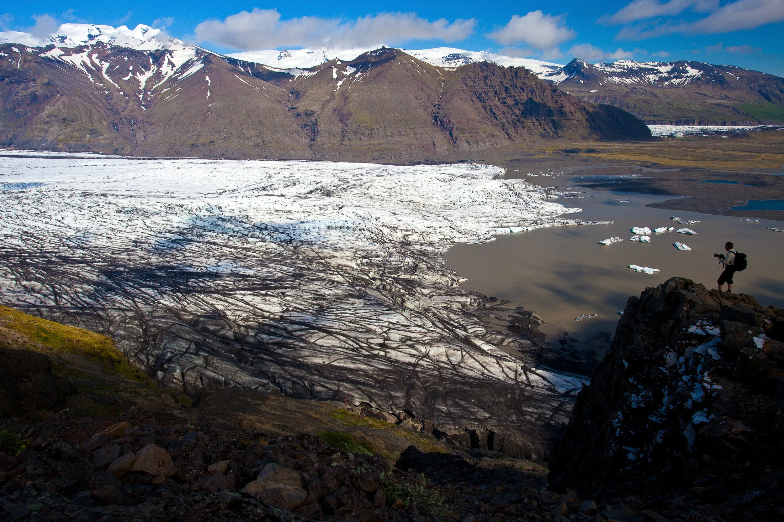 Skaftafellsjökull, Iceland — a lone photographer stands above the glacier tongue, where ash-dark crevasses spill into a milky proglacial lagoon beneath snow-dusted peaks.