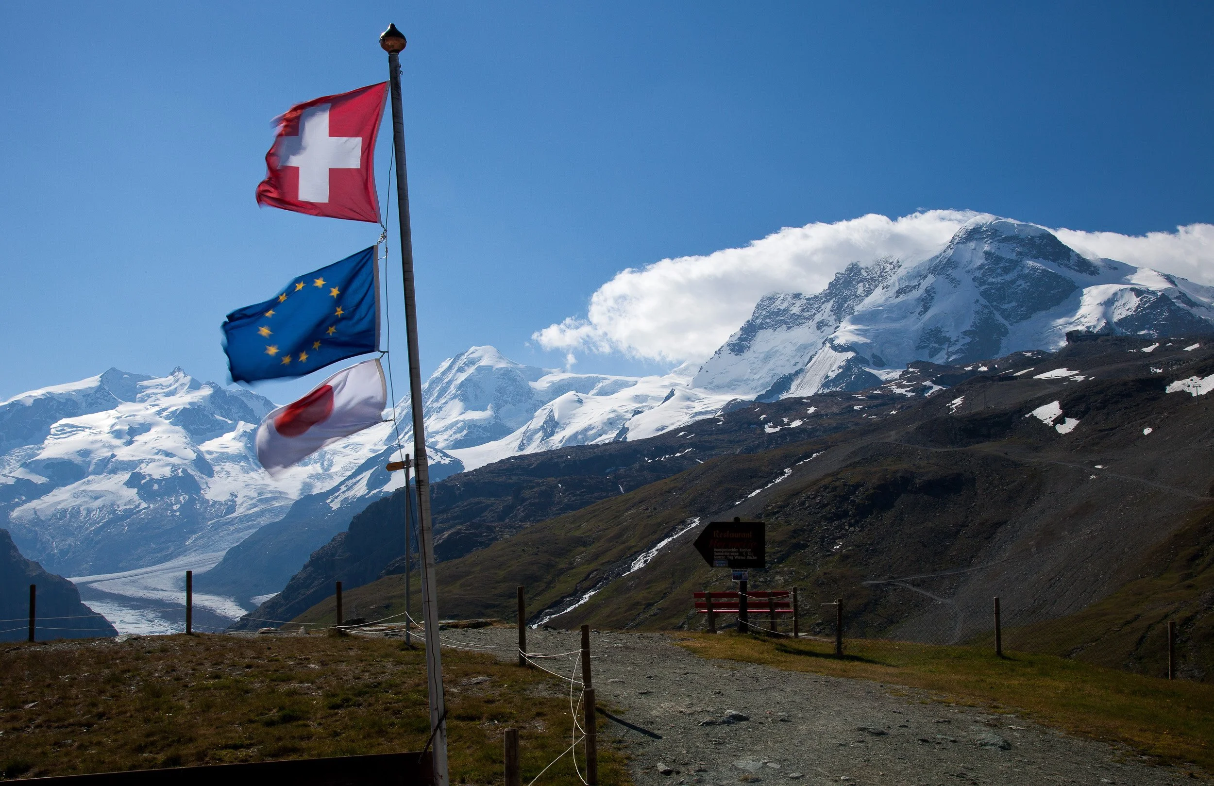 Swiss, European and Valais flags fluttering in front of snow-covered peaks and a long glacier above Zermatt.