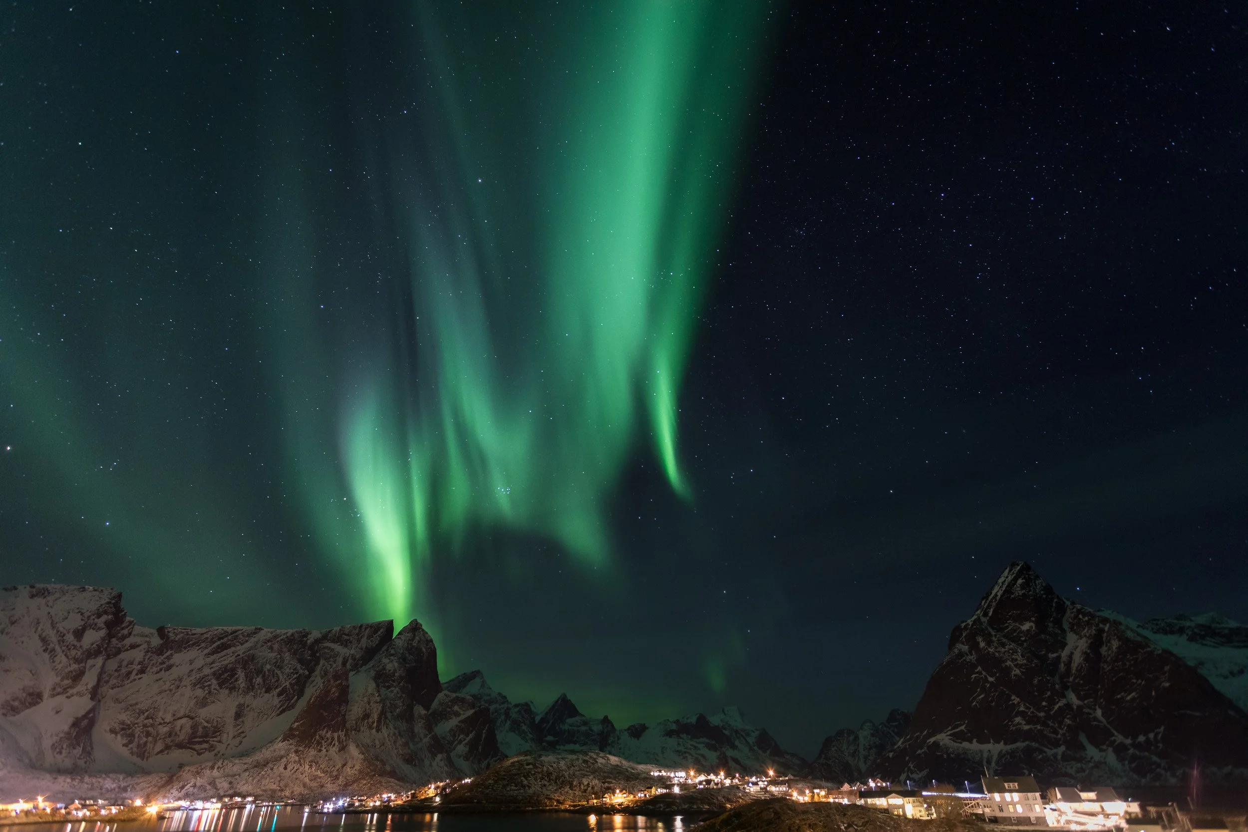 Northern Lights above Reine in the Lofoten Islands – tall curtains of green aurora rise behind jagged mountain peaks and a small Arctic village, showcasing some of Norway’s most spectacular Northern Lights scenery.