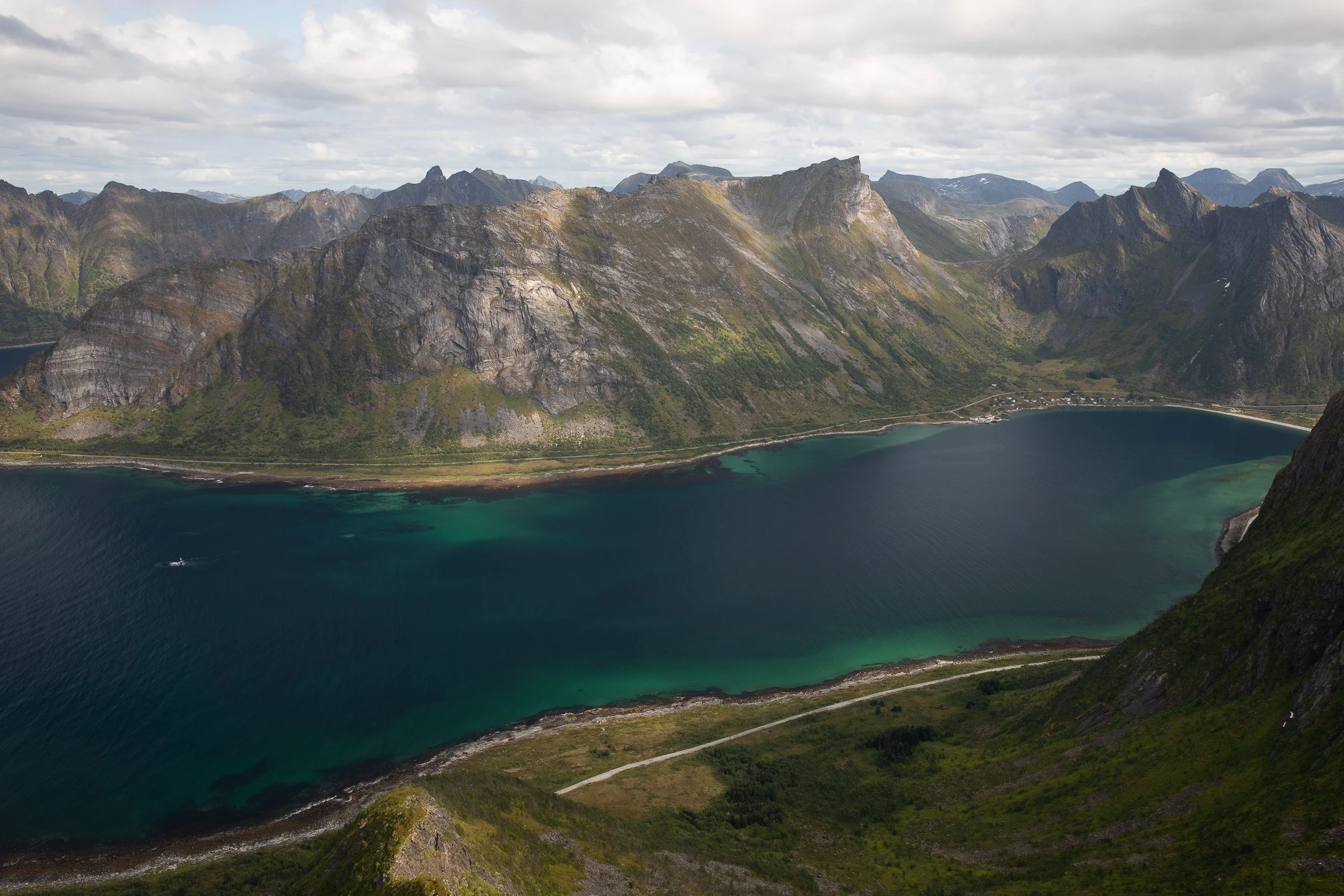 A wide view from Husfjellet across turquoise fjord water, steep mountain walls and shifting cloud light on the Senja coast.