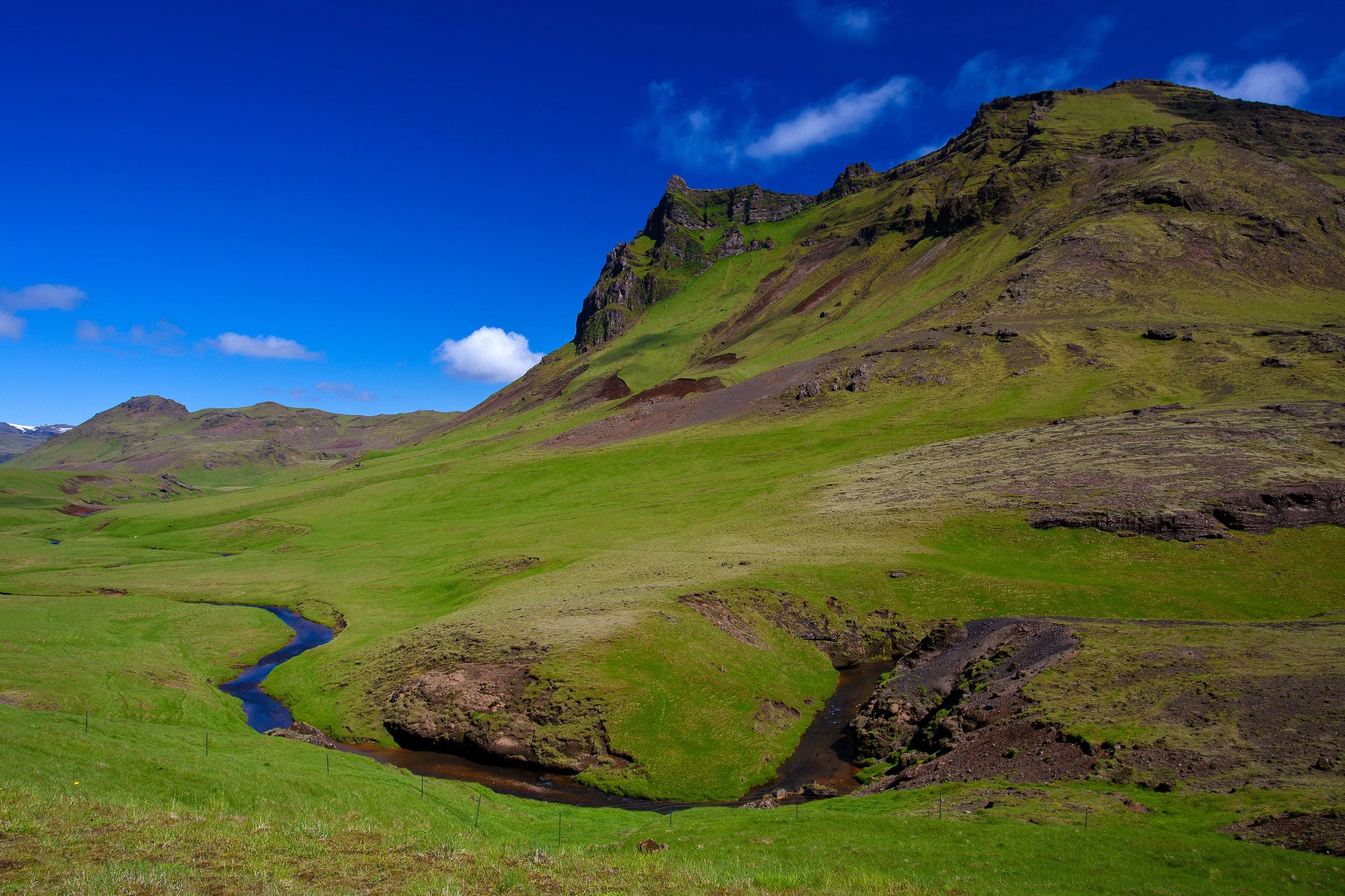 A winding river ribboning through vivid green slopes, where Iceland’s volcanic terrain softens into open, rolling countryside.