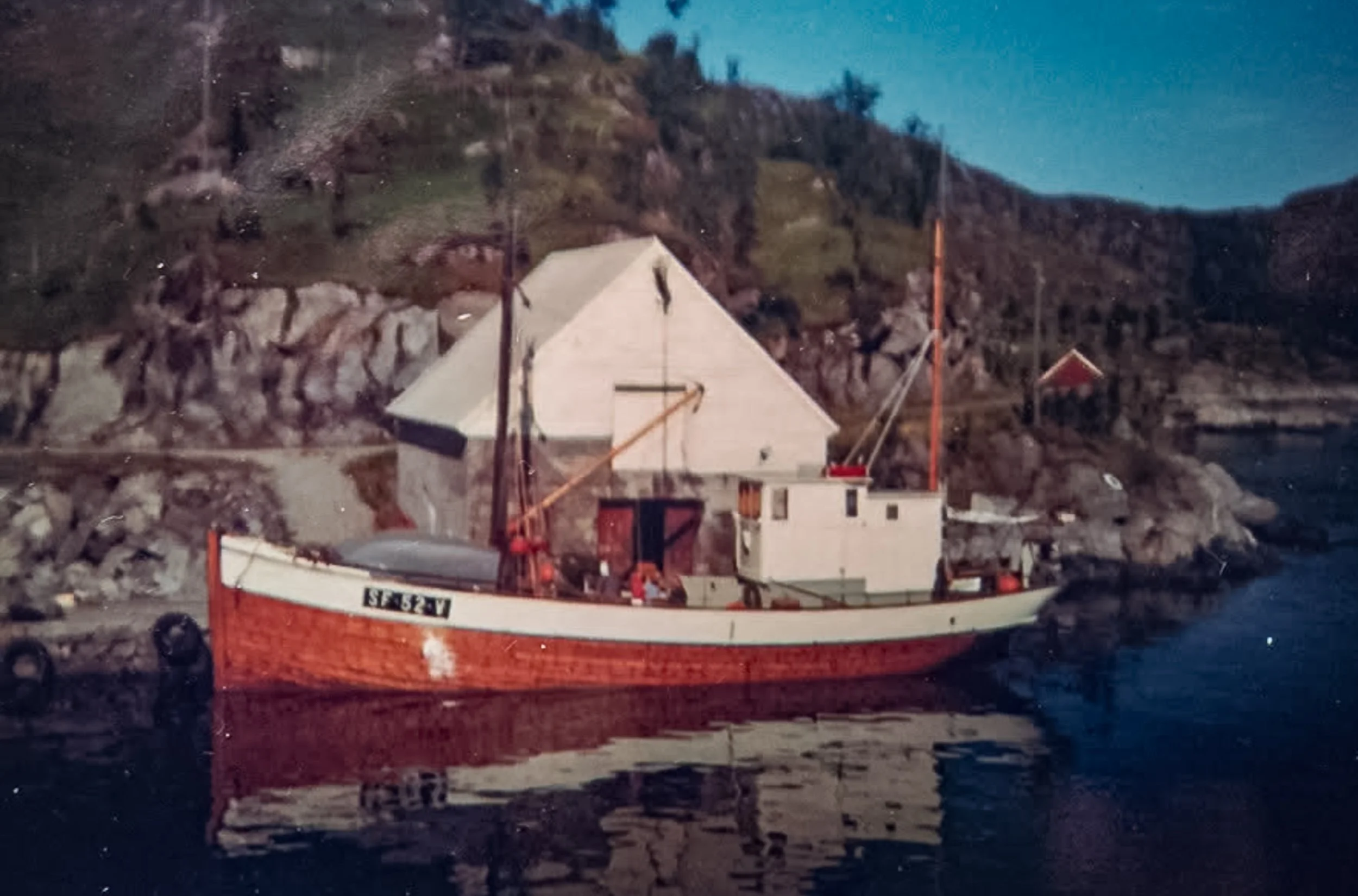 Motorkutteren «Hovdenes» SF-52-V, med rødt skrog og hvitt overbygg, fortøyd ved sjøbua i Vågen i Husevåg.