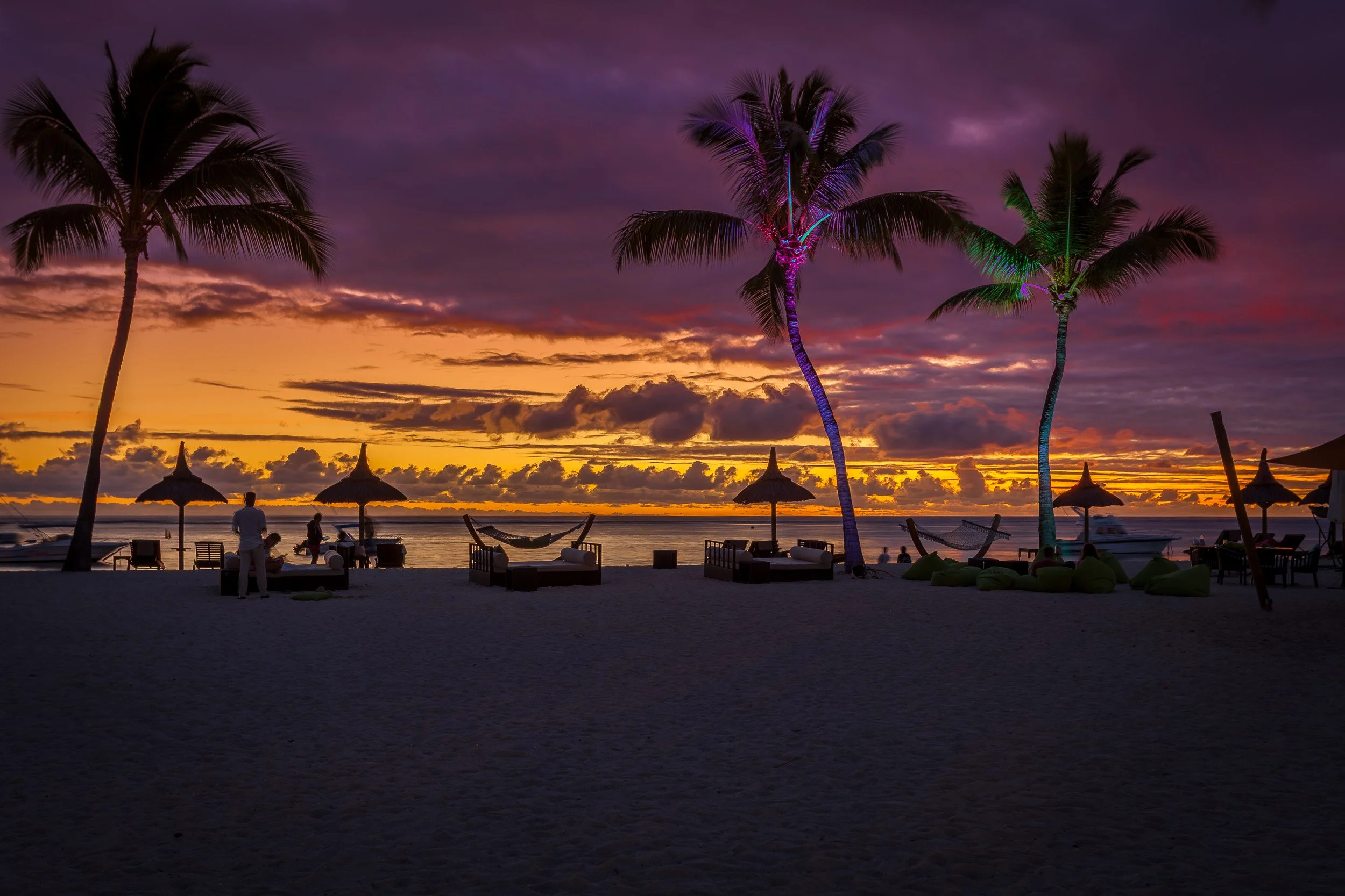 Dramatic sunset over Flic en Flac beach on Mauritius, with glowing purple clouds, lit palm trees and daybeds along the tranquil lagoon.