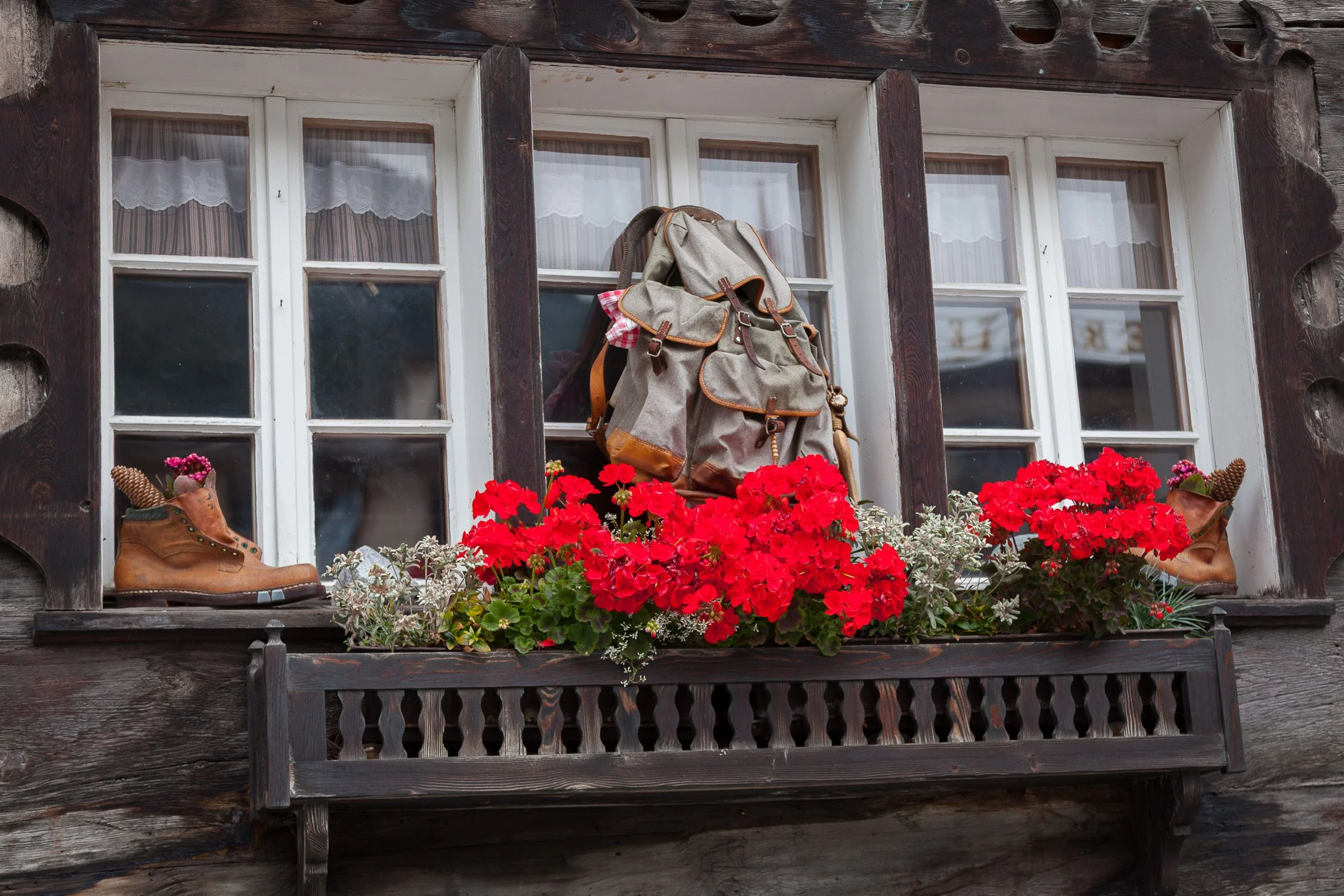 Old wooden house window in Zermatt decorated with hiking boots used as flower pots, a vintage rucksack and bright red geraniums.