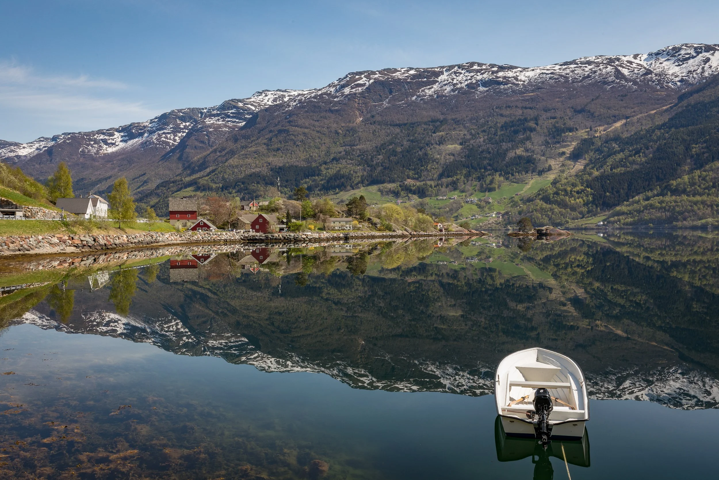 A small boat at rest on a mirror-calm fjord, with red boathouses and mountains reflected in the water.