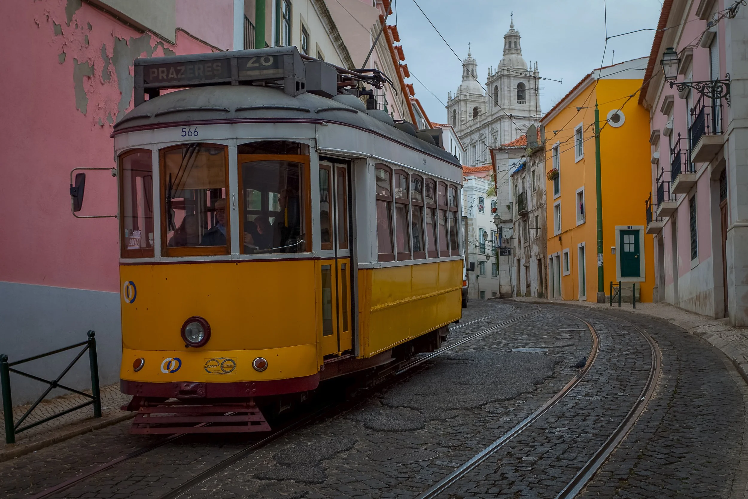 A yellow Lisbon tram follows the narrow tracks through Alfama, framed by coloured façades and the white towers of São Vicente de Fora.