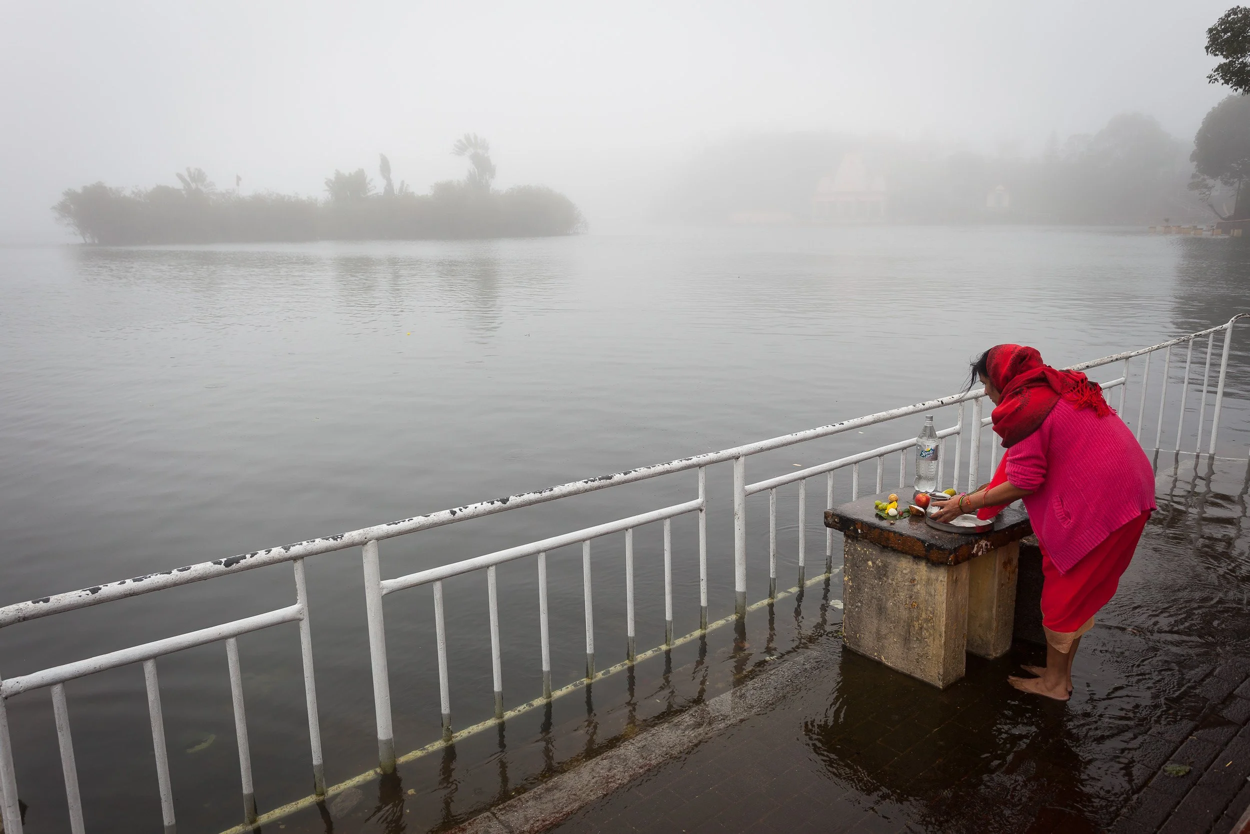 Hindu devotee in red making fruit offerings at the mist-covered Ganga Talao crater lake in the highlands of Mauritius.