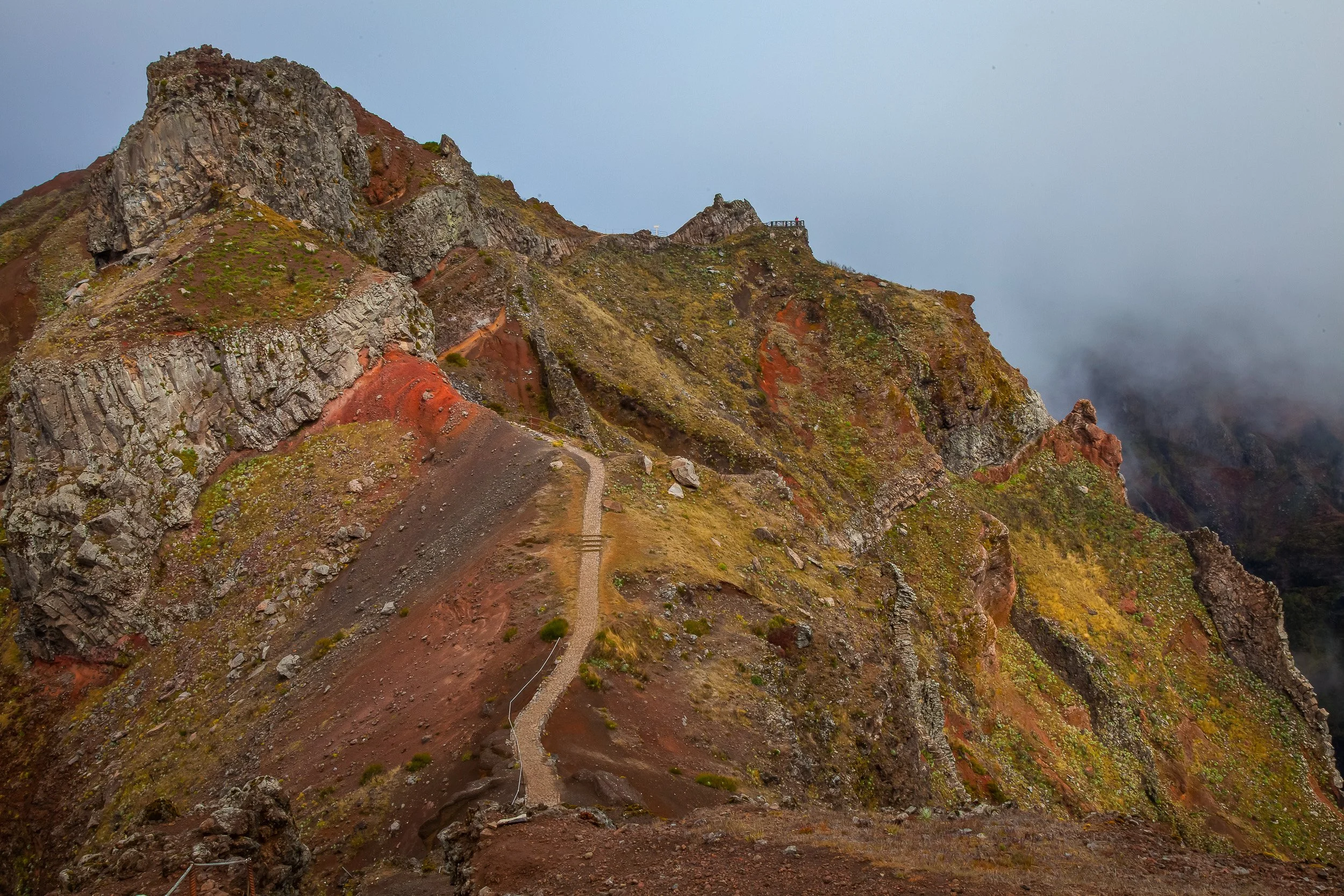 The ridge below Pico do Arieiro, where a narrow mountain path cuts across red and grey rock before disappearing towards cloud-covered peaks.