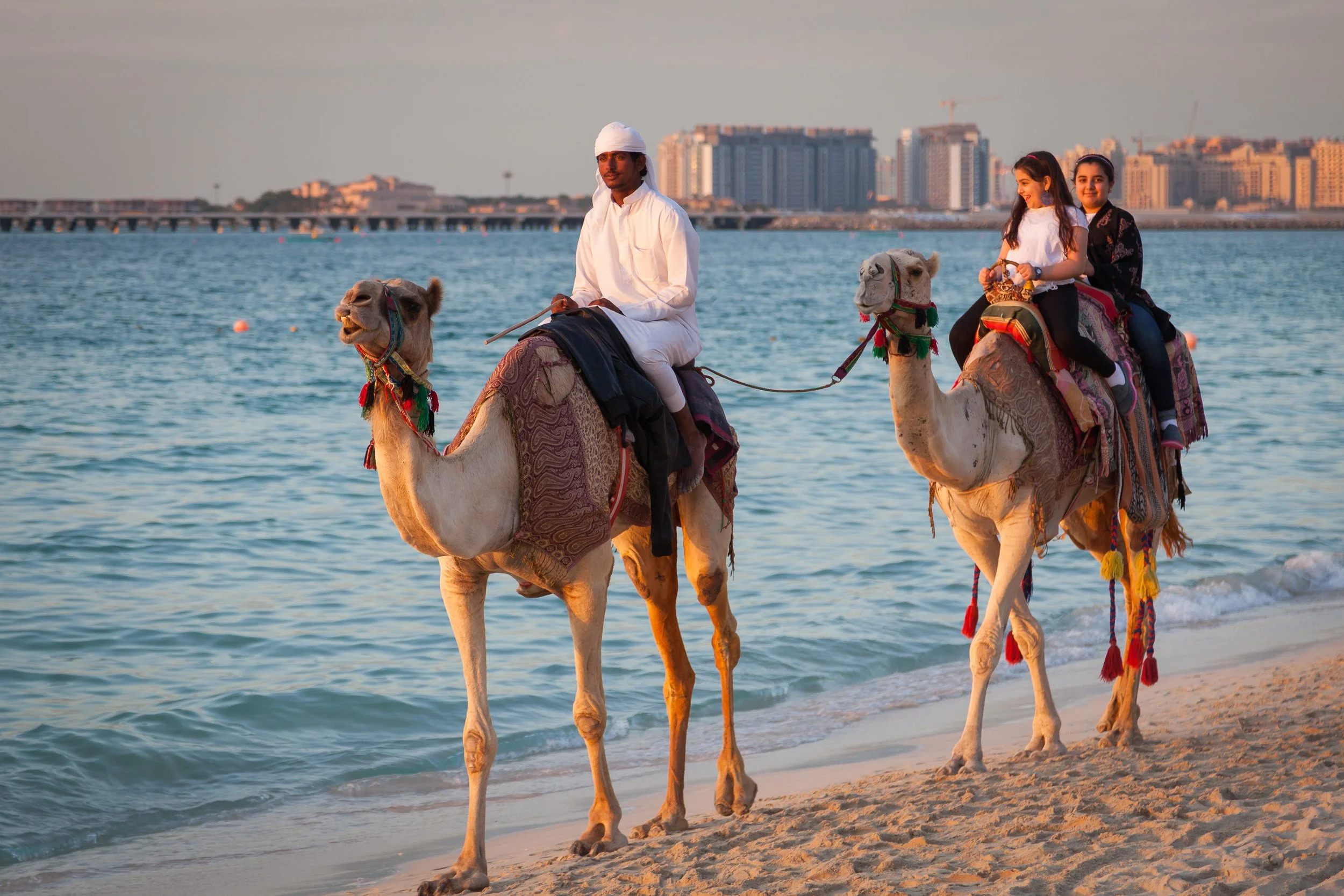 Camel riders along the shoreline at golden hour, with Dubai’s waterfront skyline rising in the distance.