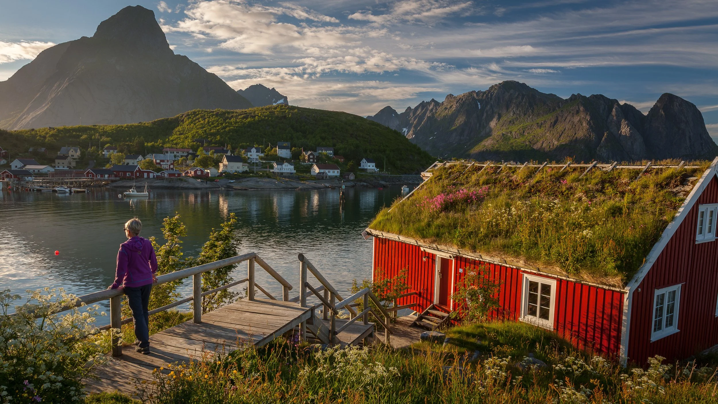 Reine in the Lofoten Islands, Norway – harbour, boats and traditional rorbuer nestled between sharp mountain walls and a calm fjord. A woman stands on the shore looking towards the midnight sun on the horizon, while reflections of red cabins and fish