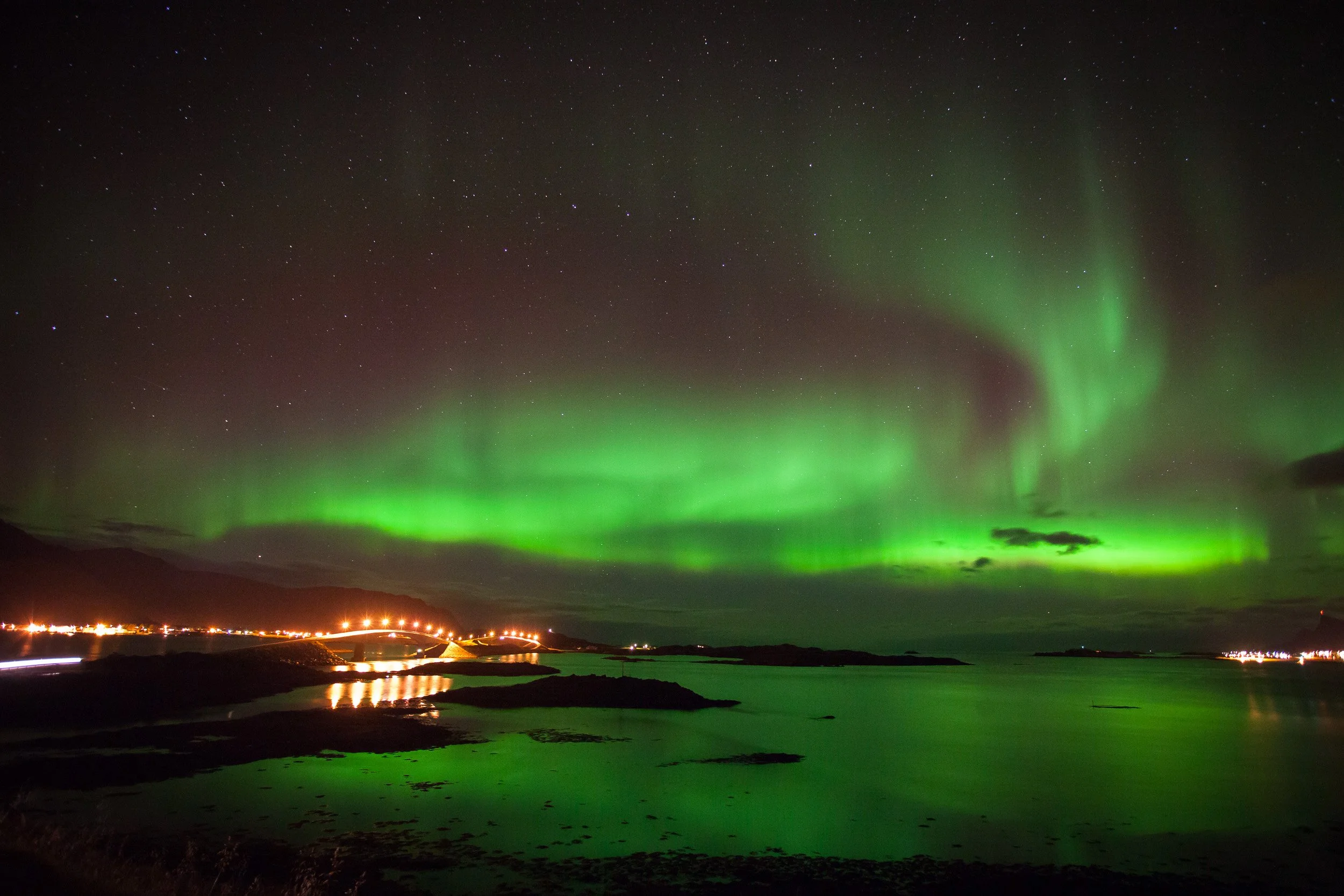 The northern lights sweep across the sky above Fredvang in Lofoten, while the warm road lights on the Flakstad bridges trace a golden line beneath the green aurora.