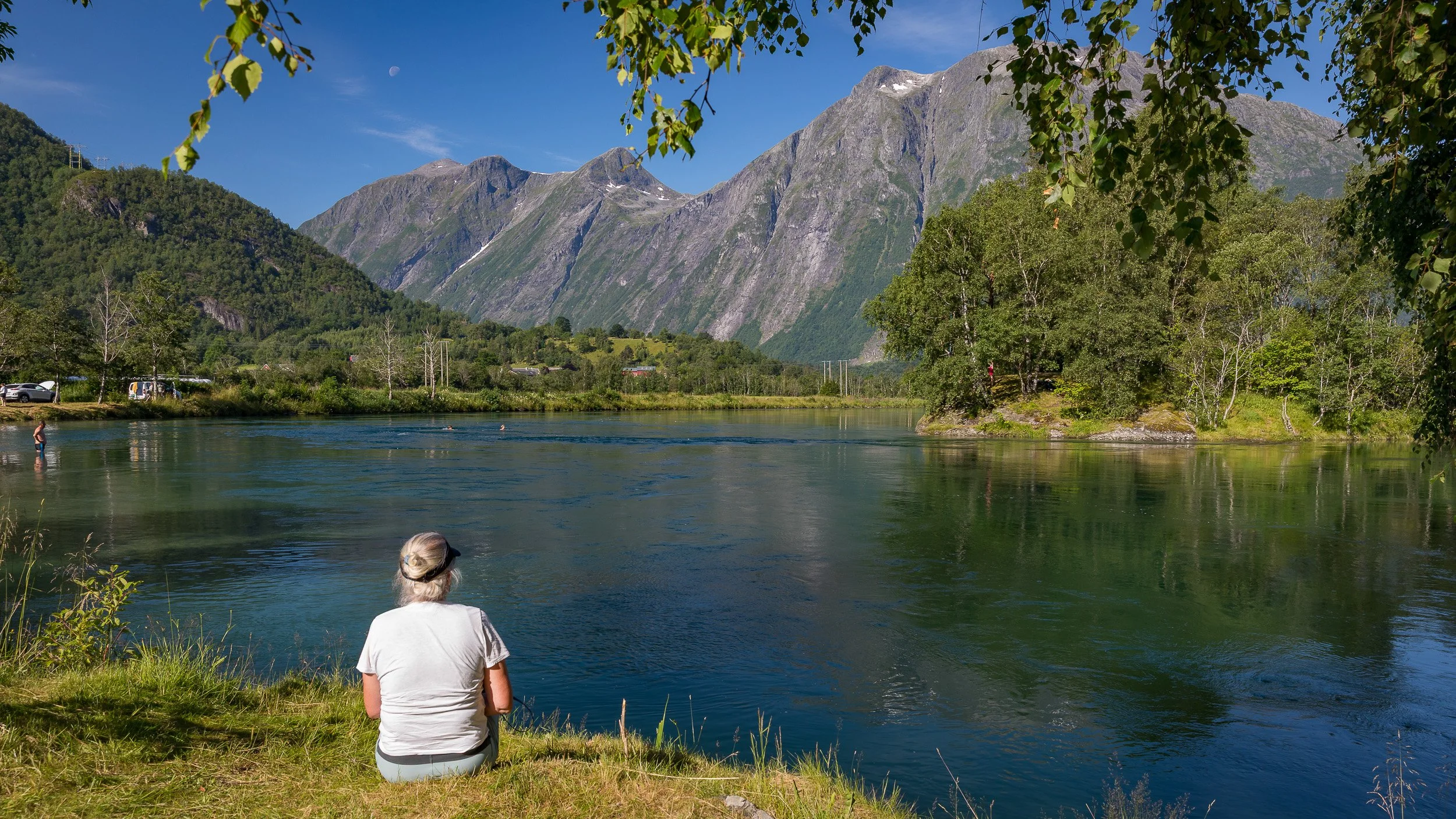 Åndalsnes, Norway – a fjord town in the Romsdalen region, surrounded by steep mountains and deep valleys. A woman sits by the riverbank looking towards the high peaks in the background, adding a quiet human presence to one of Norway’s most dramatic i