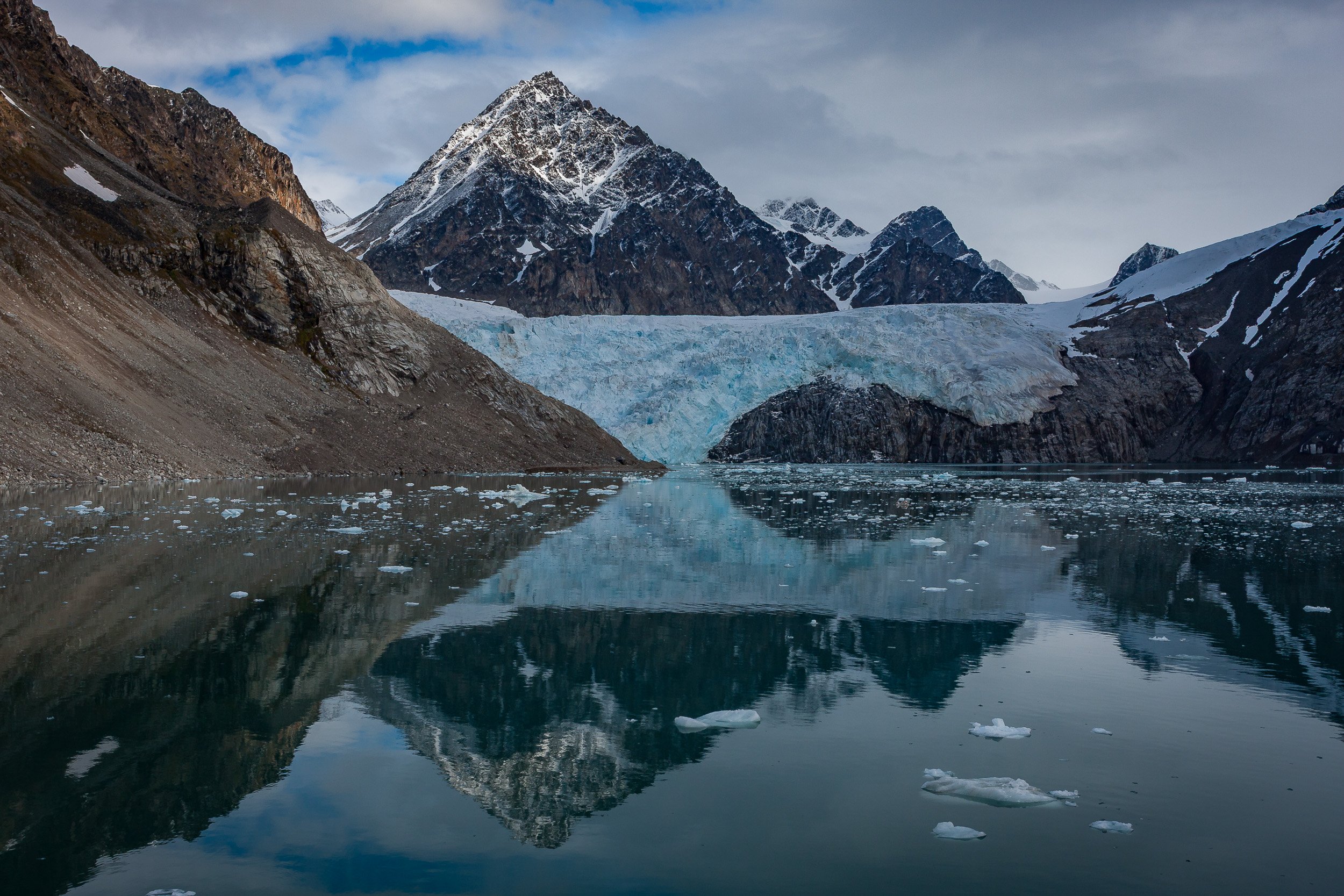 Glacier ice and steep rock walls enclose a still lagoon, where mountain shapes are repeated in the water.