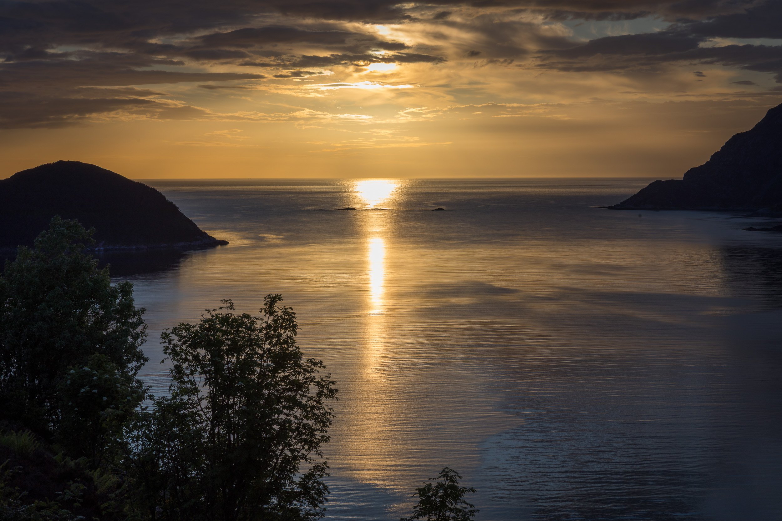 Midtsommerkveld ved utløpet av Nordfjord, sett fra Husevågøy, med solen på vei ned mot havet og gyllent lys over fjord, kystfjell og åpen horisont.