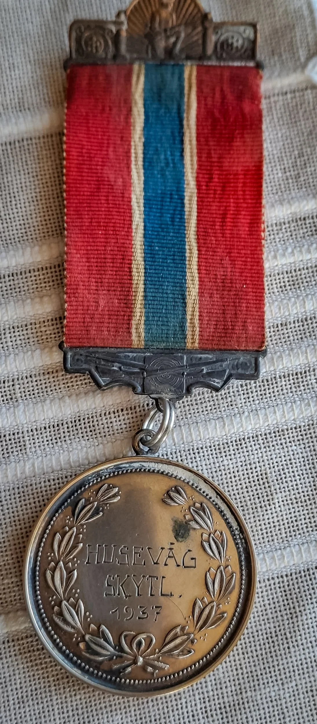 Medalje fra Husevåg skytterlag med rødt bånd og blå midtstripe, merket «HUSEVÅG SKYTL. 1937».
