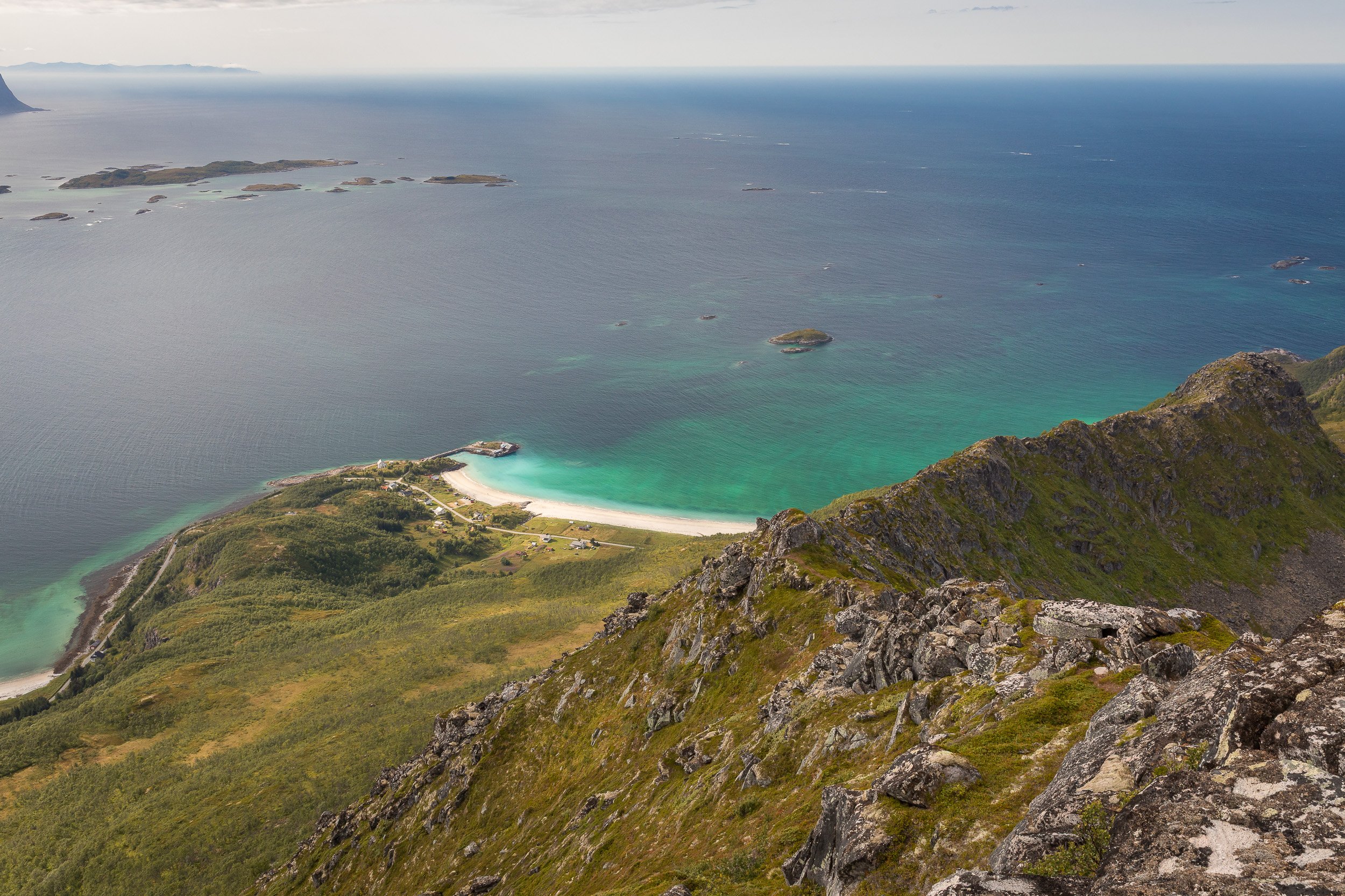 From Husfjellet, a white beach and clear turquoise water appear far below, contrasting with the rough mountain ridge in the foreground.