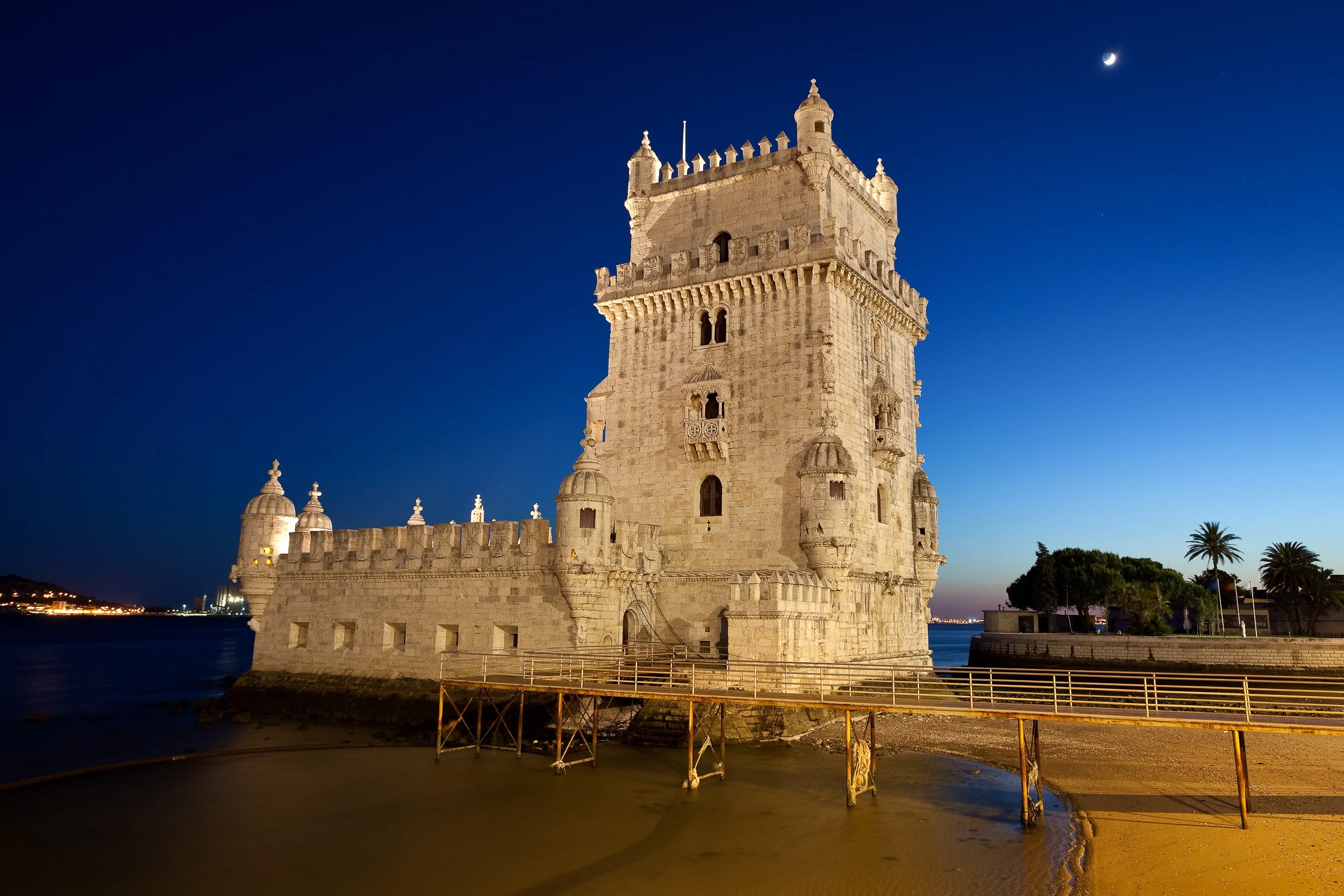 Belém Tower at blue hour, illuminated beside the Tagus River with a crescent moon above the darkening sky.