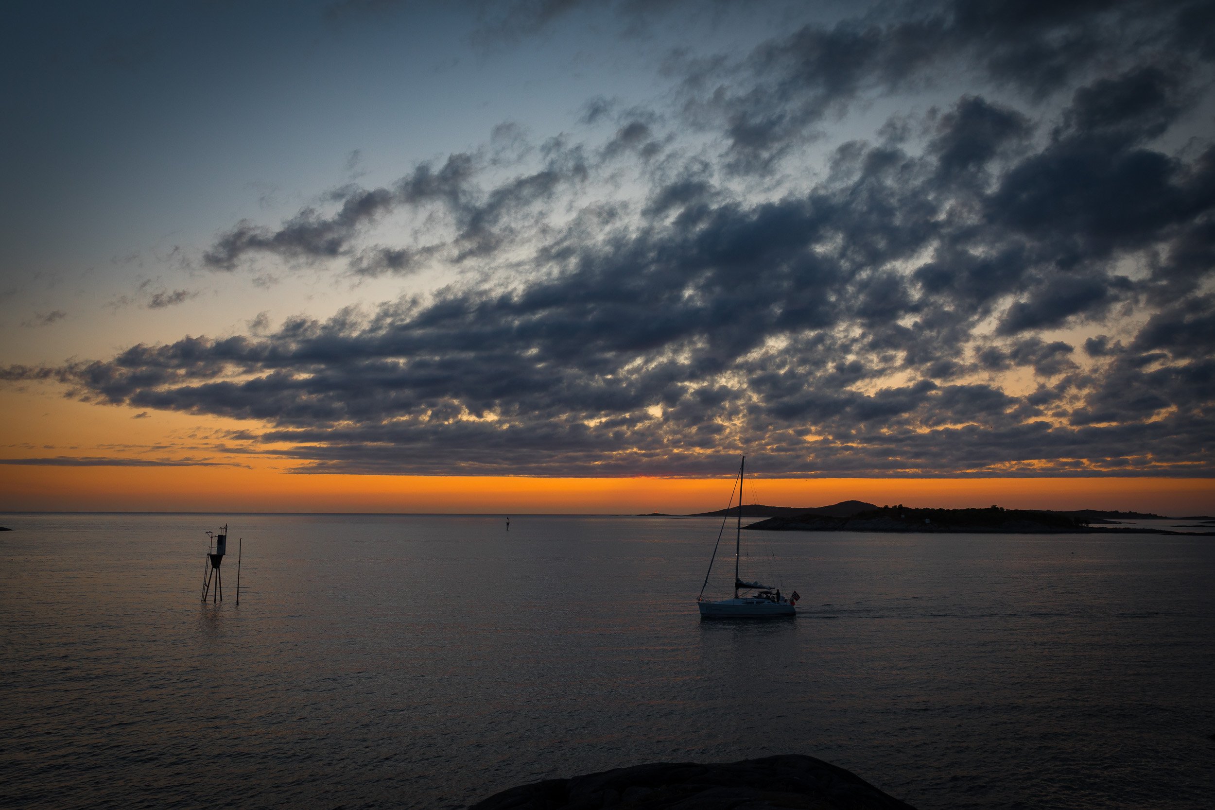 A sailboat moves through the quiet water at Hamn as dark clouds gather above the orange glow of a late Senja sunset.