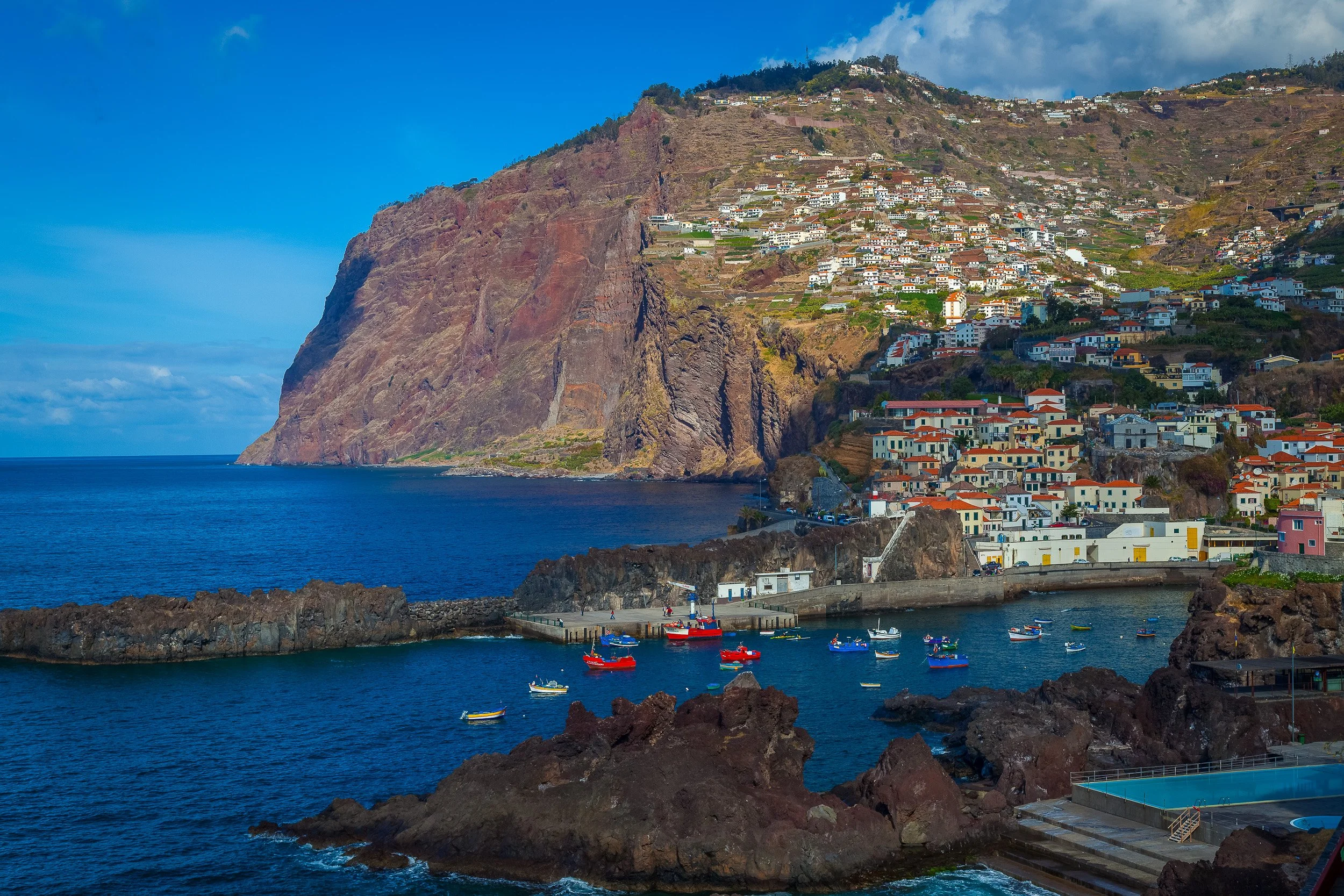 Câmara de Lobos seen from above, with fishing boats in the harbour below steep cliffs and white houses climbing the hillside towards the sunlit ridge.
