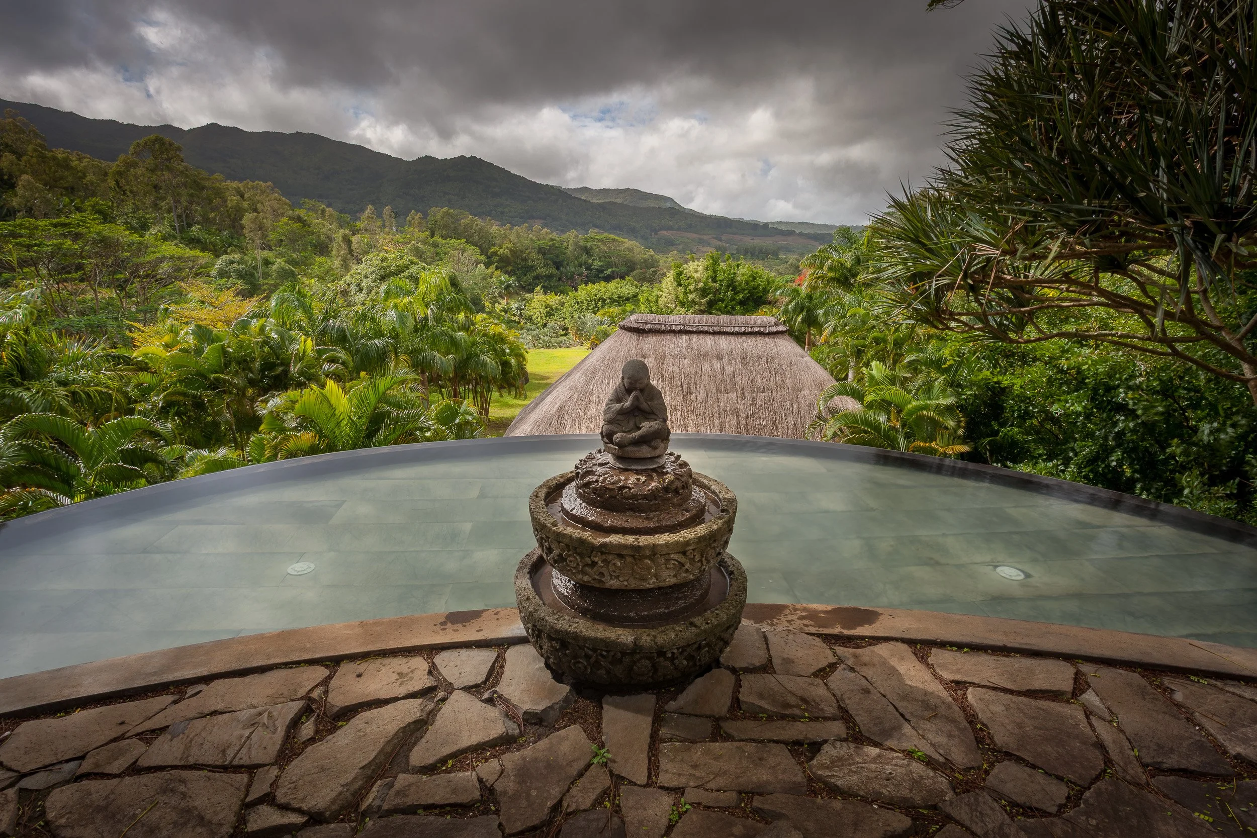 Stone meditating figure above a curved infinity pool at Lakaz Chamarel, overlooking misty green valleys in the mountains of Mauritius.