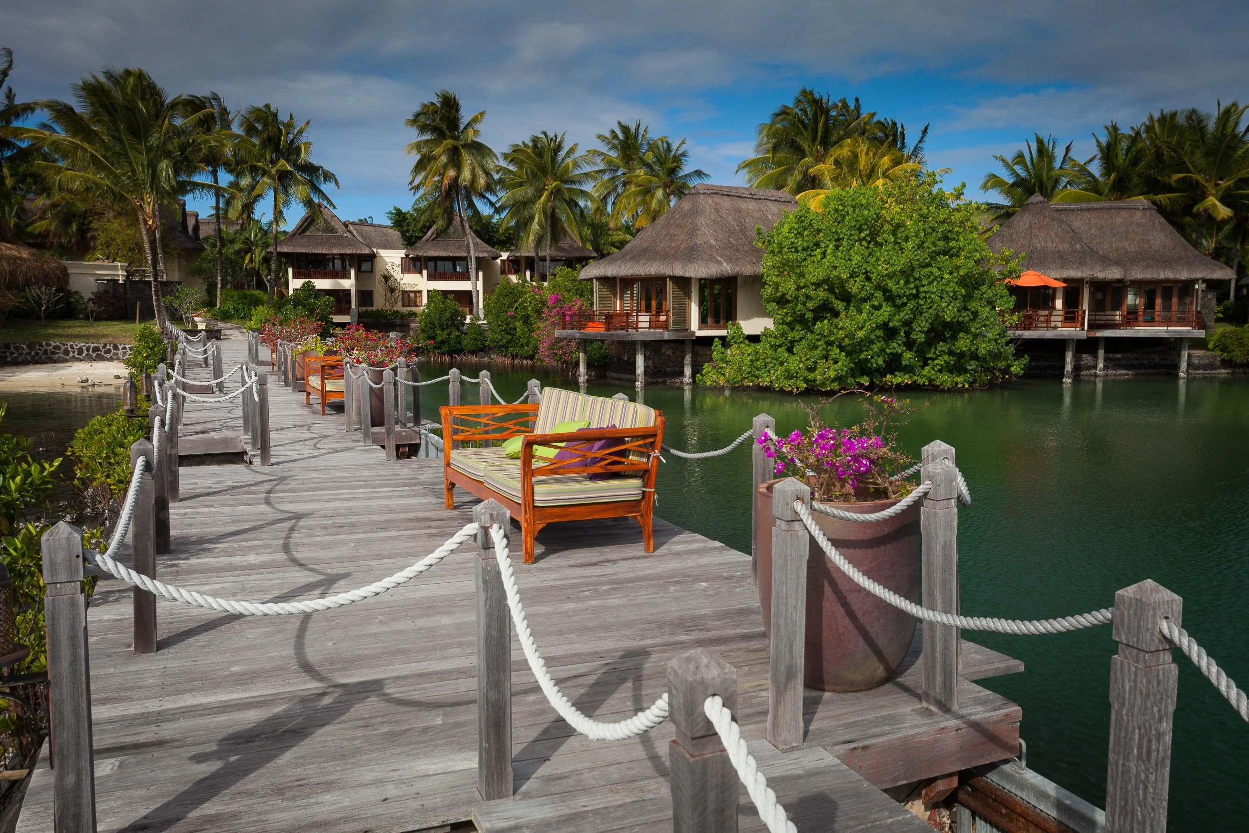 Boardwalk lined with loungers and flowering pots at Constance Le Prince Maurice, leading past over-water villas on a tranquil lagoon.