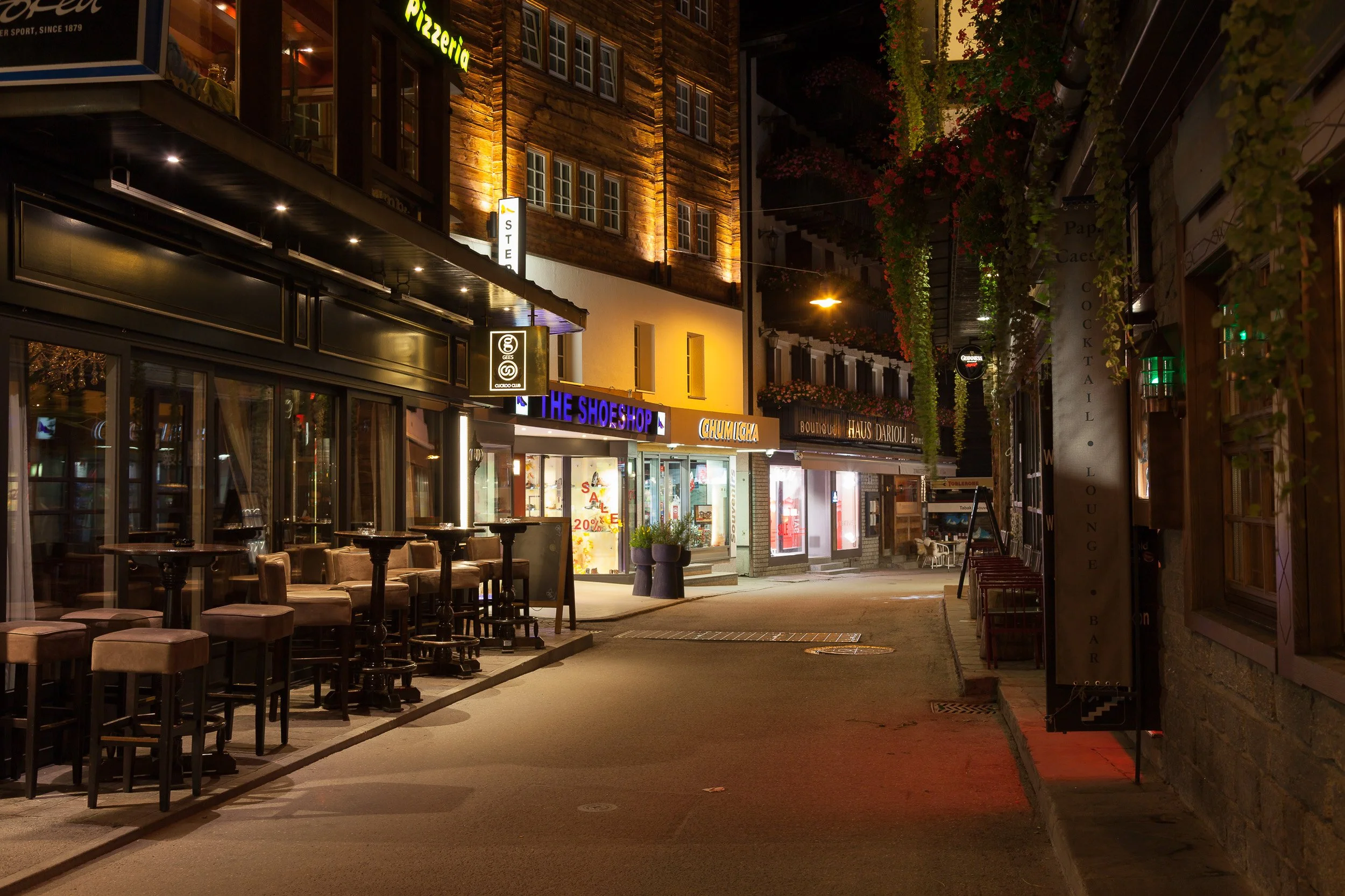 Quiet evening scene in central Zermatt: illuminated shopfronts, bars and empty café tables lining a narrow alpine street.