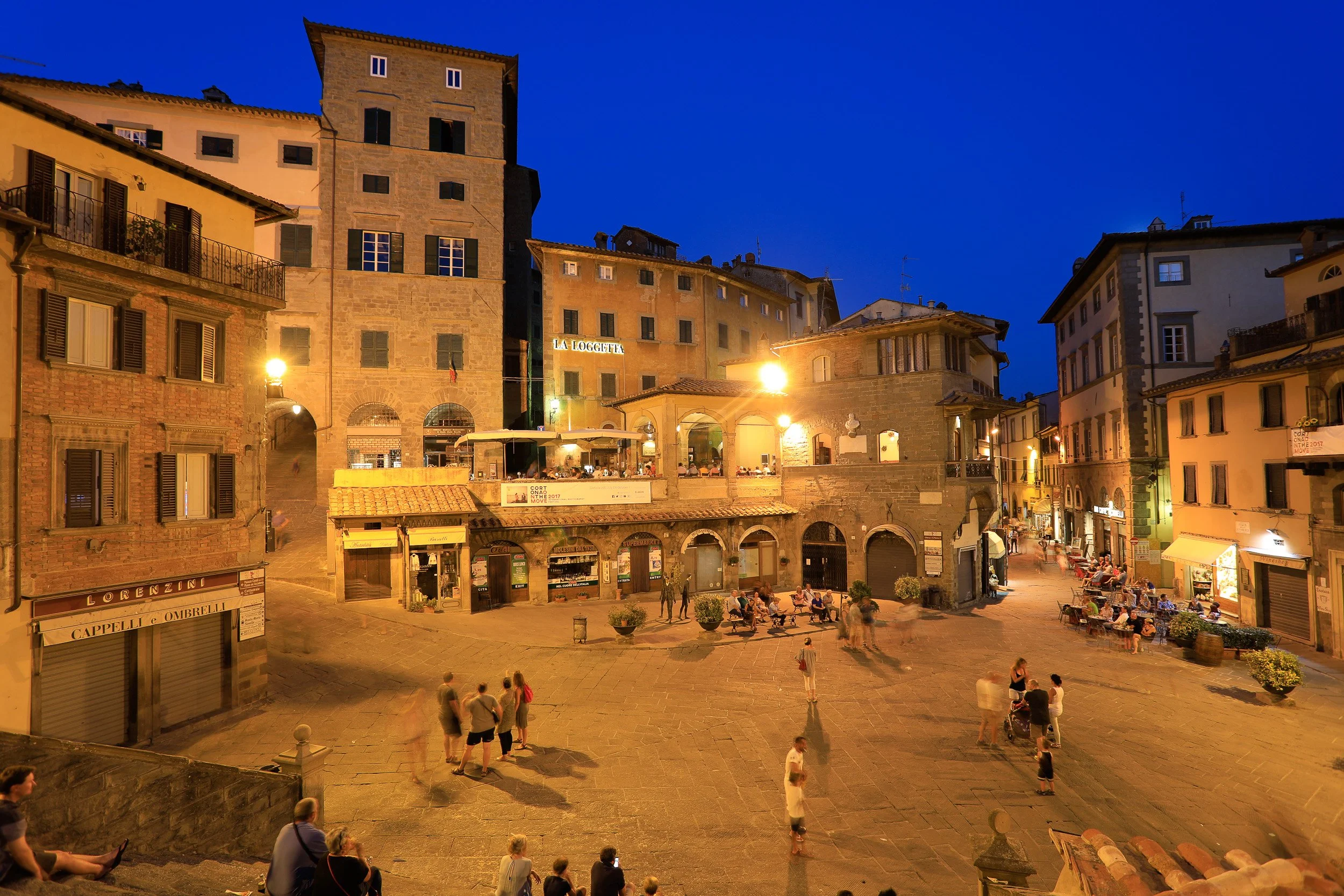 Blue hour over a Tuscan hill-town square, with warm café light and people lingering into the evening.
