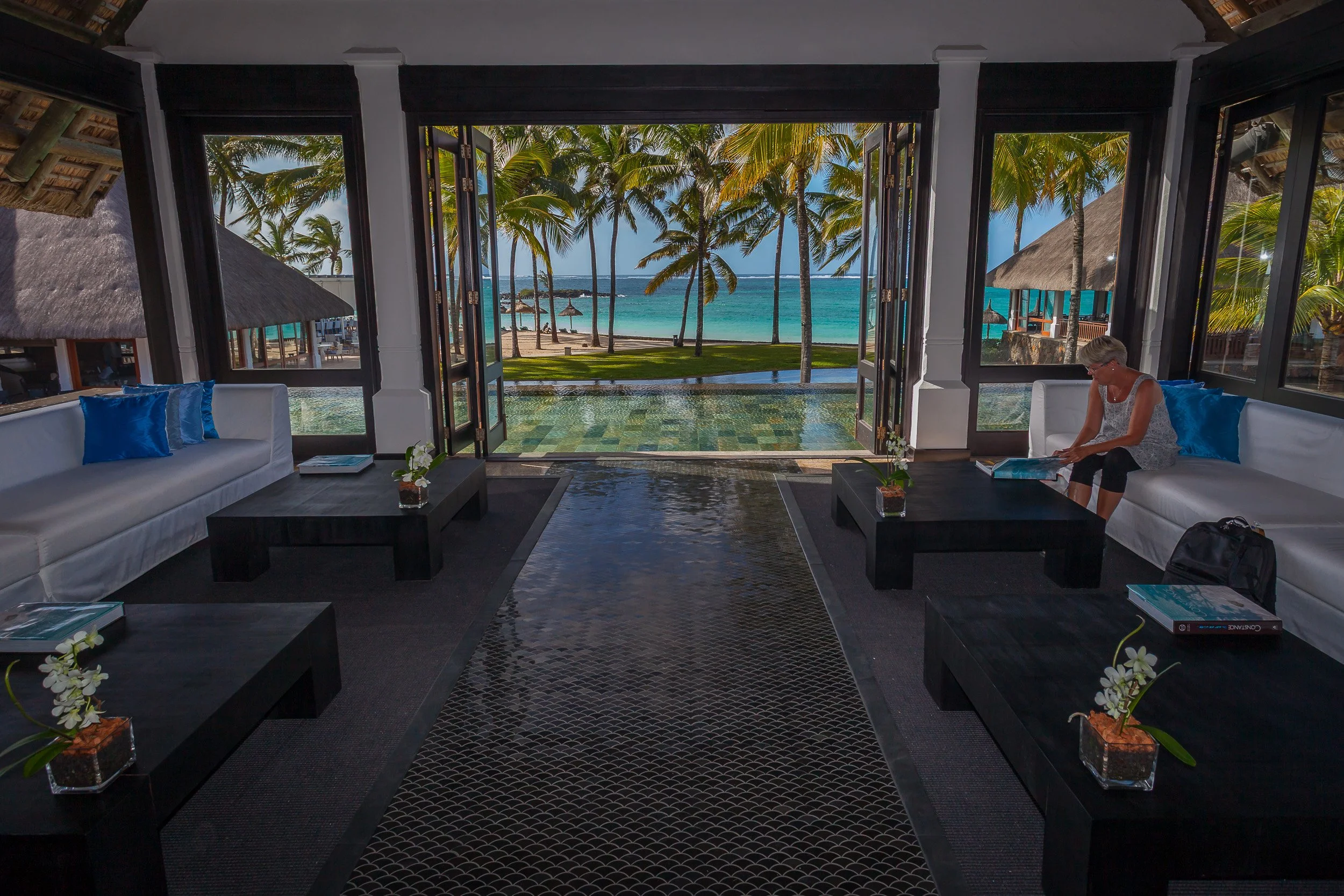 Guest relaxing with a book in the stylish reception at Constance Belle Mare Plage, looking out over pools, palms and the lagoon in Mauritius’ Flacq district.