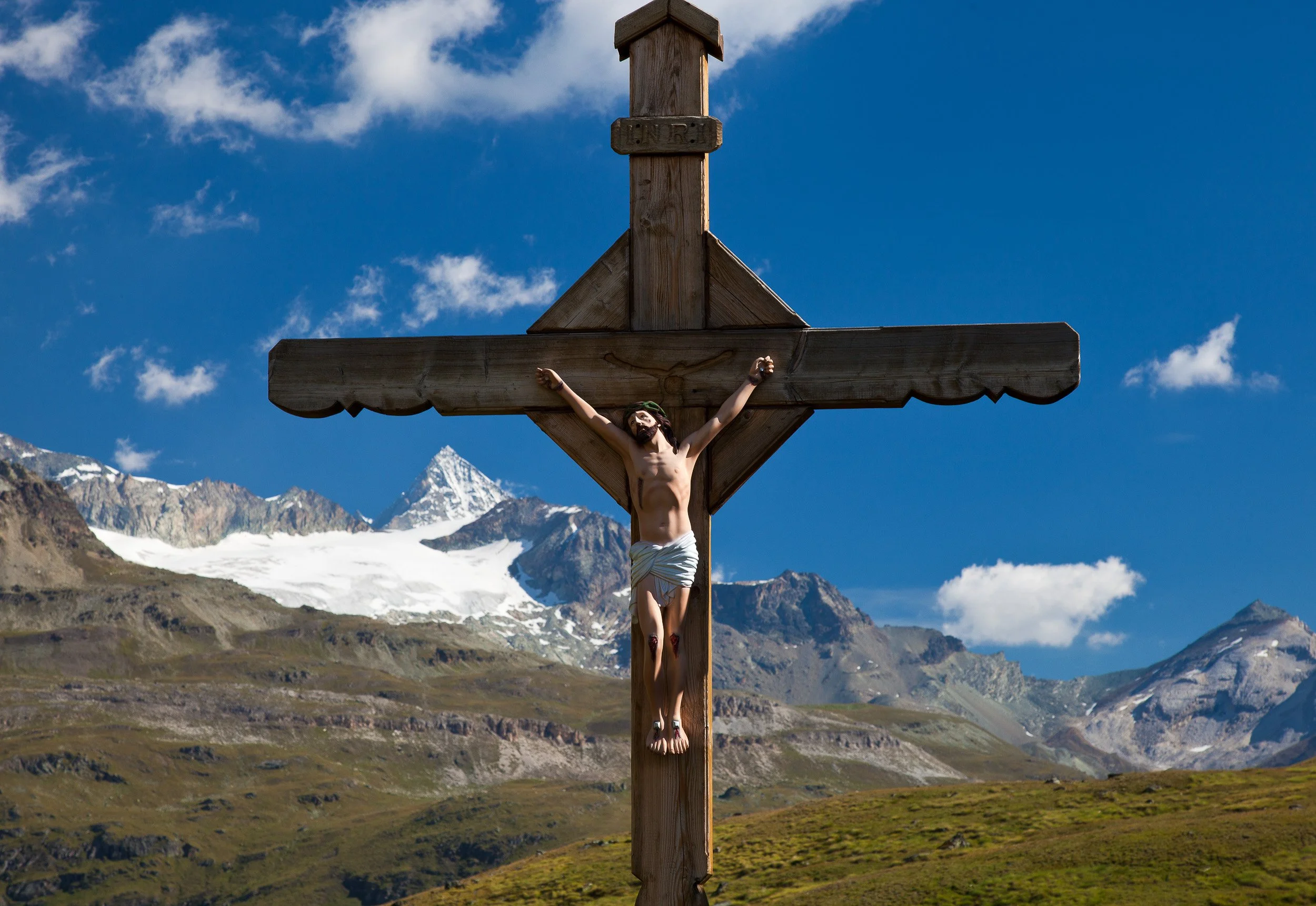 Wooden crucifix of Christ near Schwarzsee above Zermatt, framed by glaciers and rugged Swiss peaks under a clear blue sky.