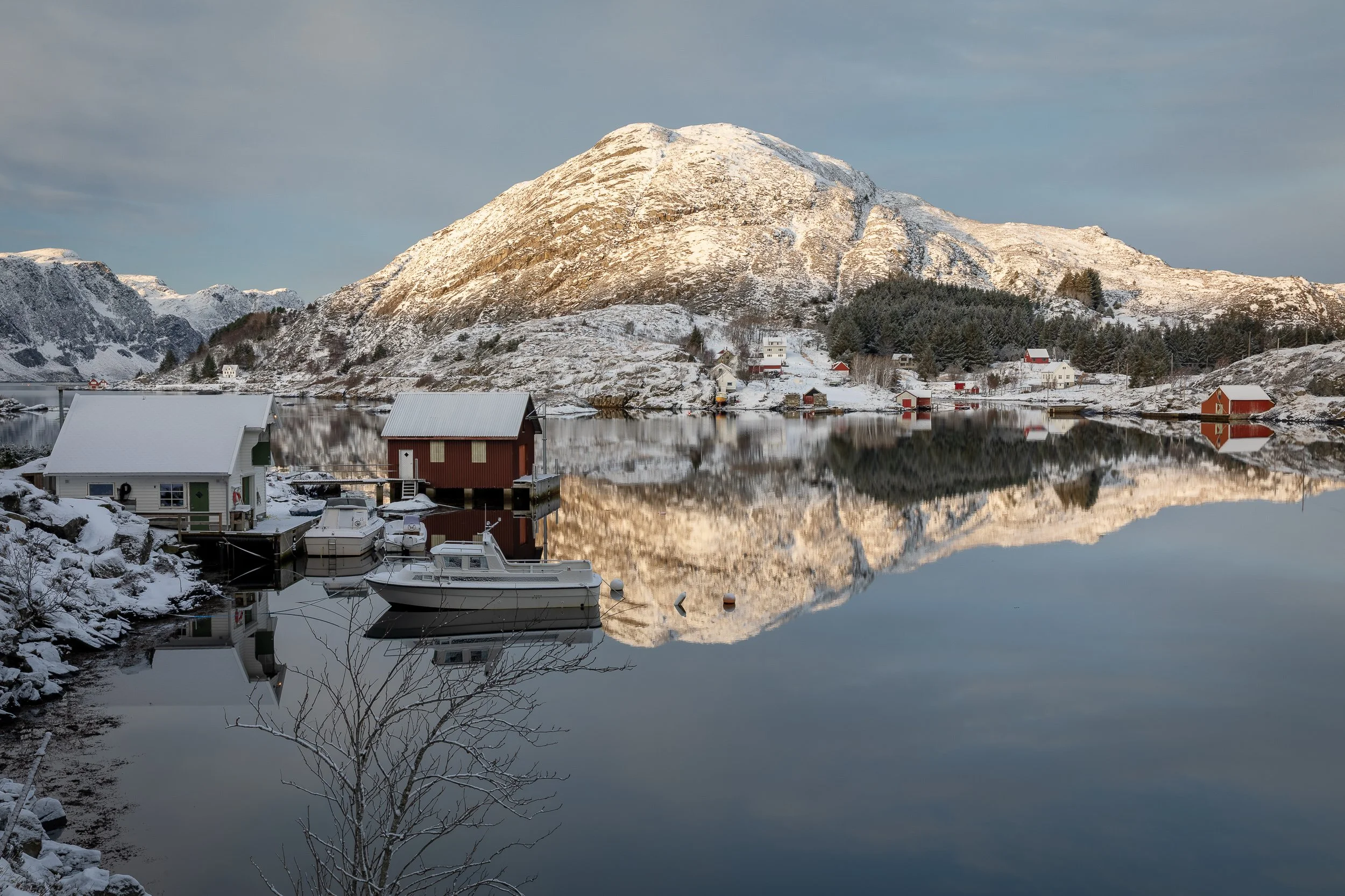 Tytingvåg på Husevågøy om vinteren, med naust, båter, snødekte fjell og speilinger i den stille sjøen.