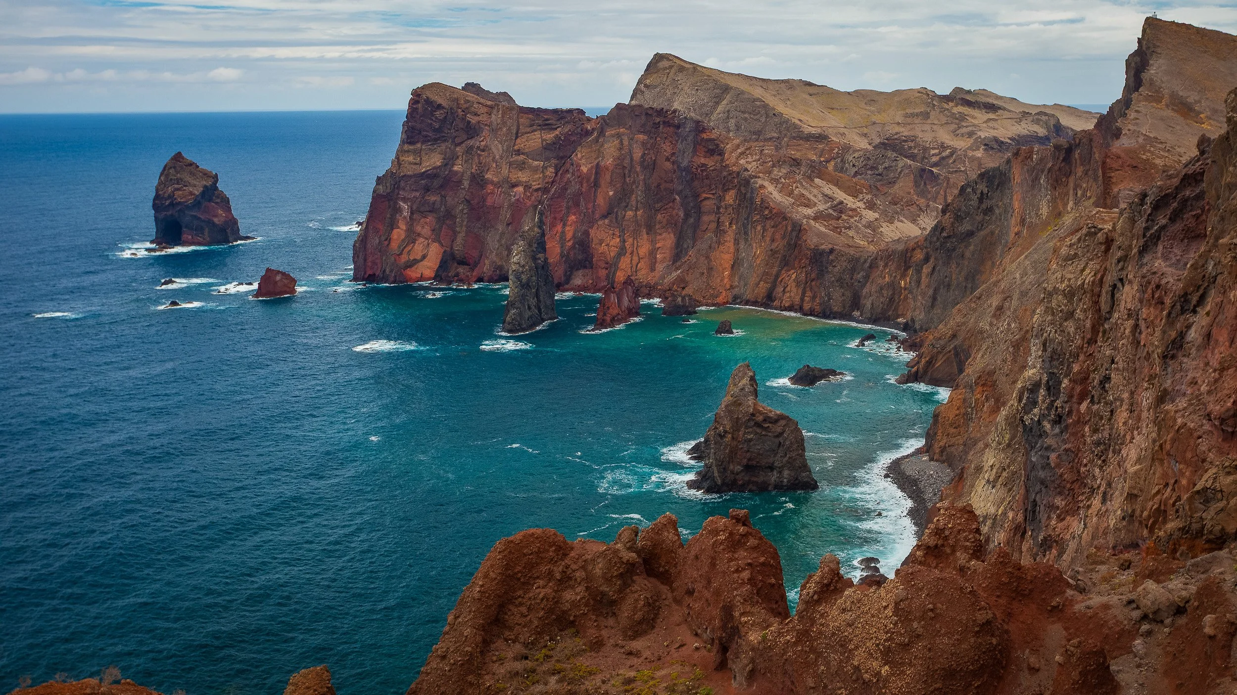 Ponta de São Lourenço in Madeira, Portugal, where red-brown volcanic cliffs and sea stacks meet turquoise Atlantic water along a raw coastal headland.