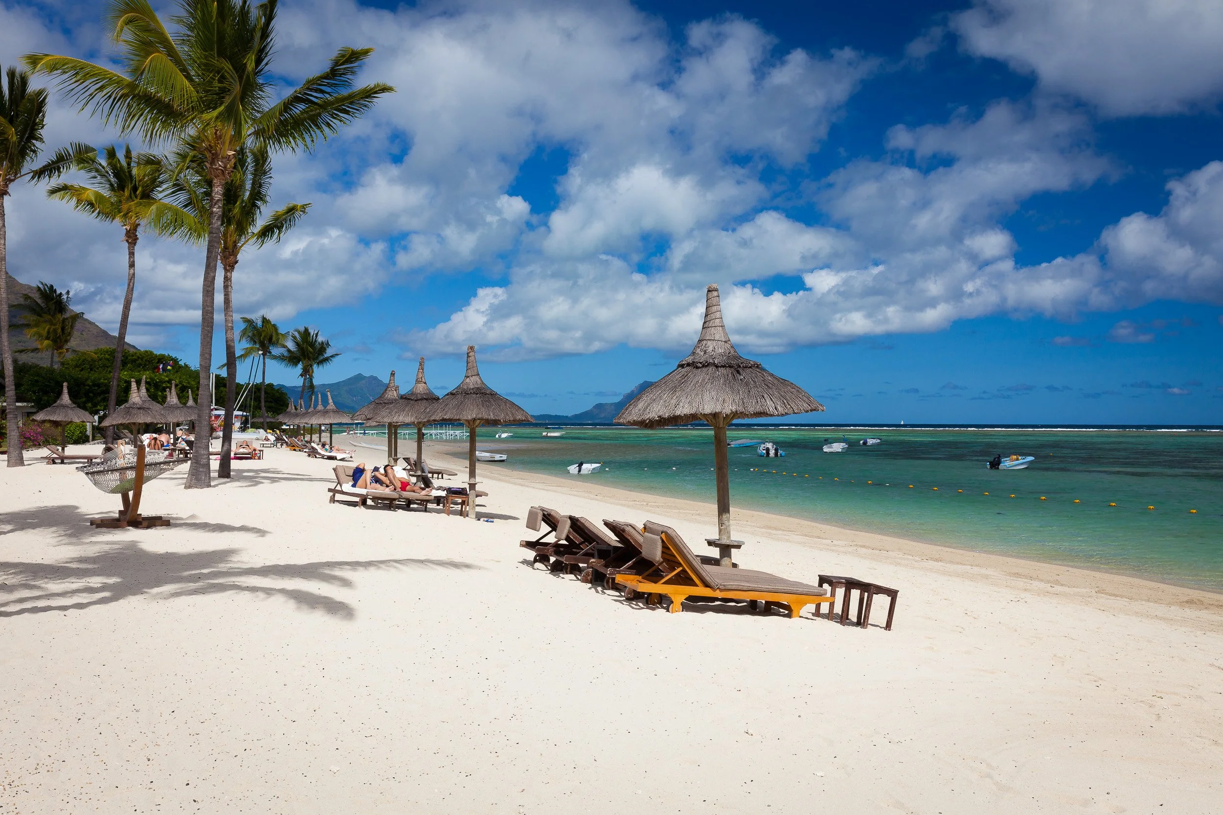 Wide sweep of white sand at Flic en Flac, Mauritius, with sun loungers under thatched parasols and green mountains framing the turquoise lagoon.