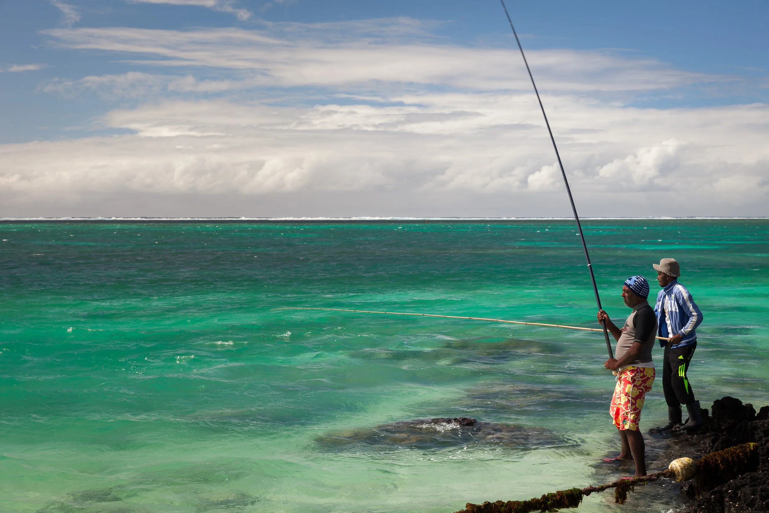 Two local fishermen standing on black rocks at Belle Mare Beach, casting long rods over the clear turquoise lagoon.