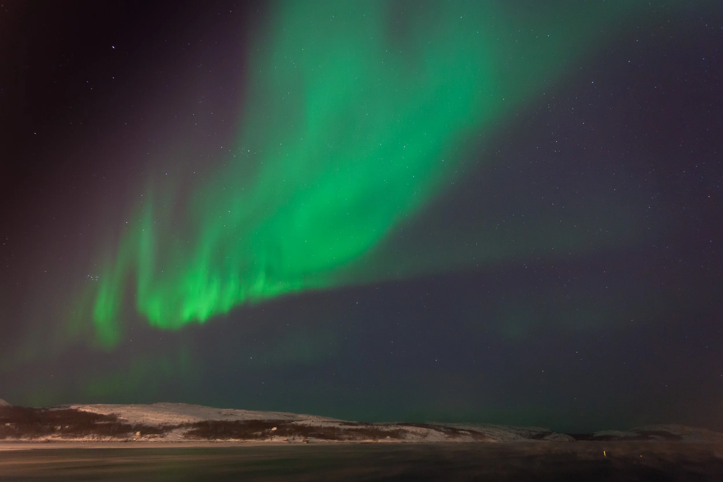 Northern lights over Kirkenes in far north-eastern Norway, with green aurora hanging above frost smoke rising from the cold fjord on a deep winter night.