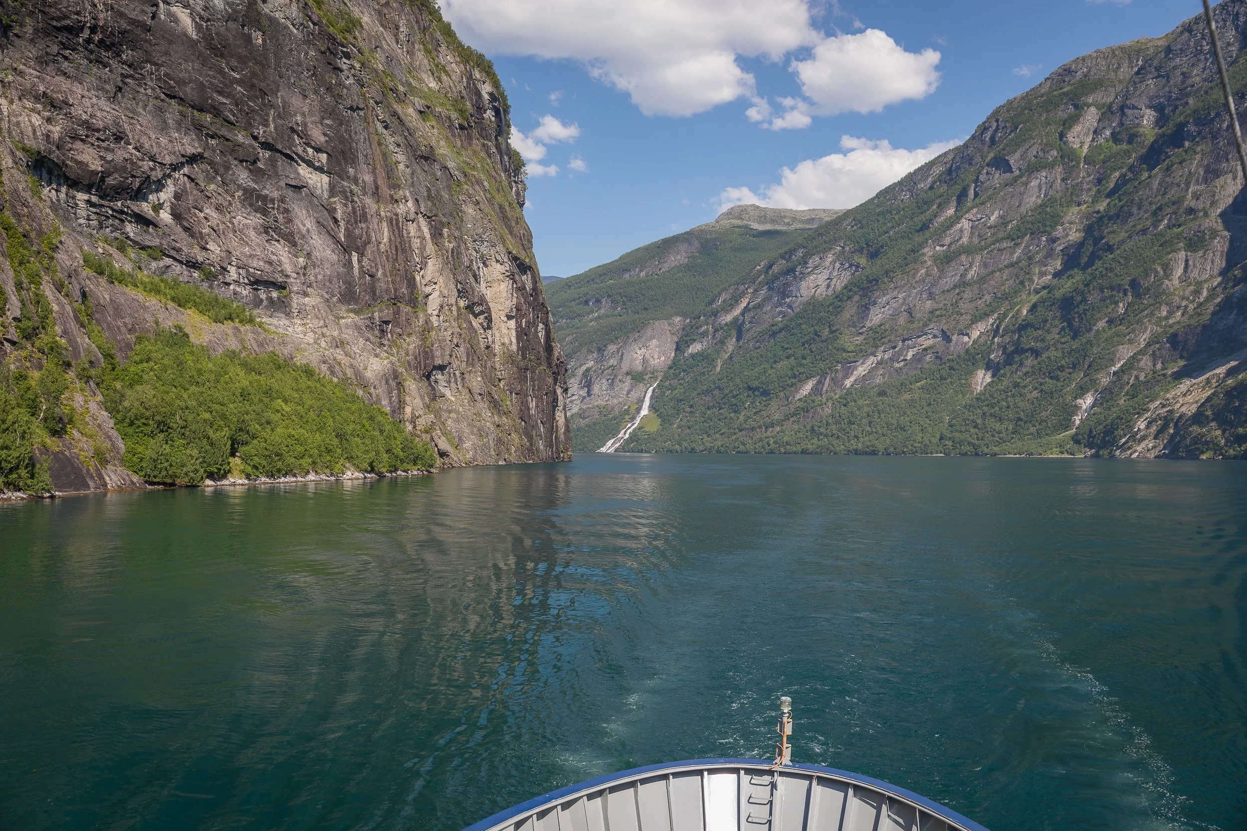 Entering Geirangerfjorden by ship — the bow points into a dramatic fjord corridor framed by cliffs and cascading waterfalls.
