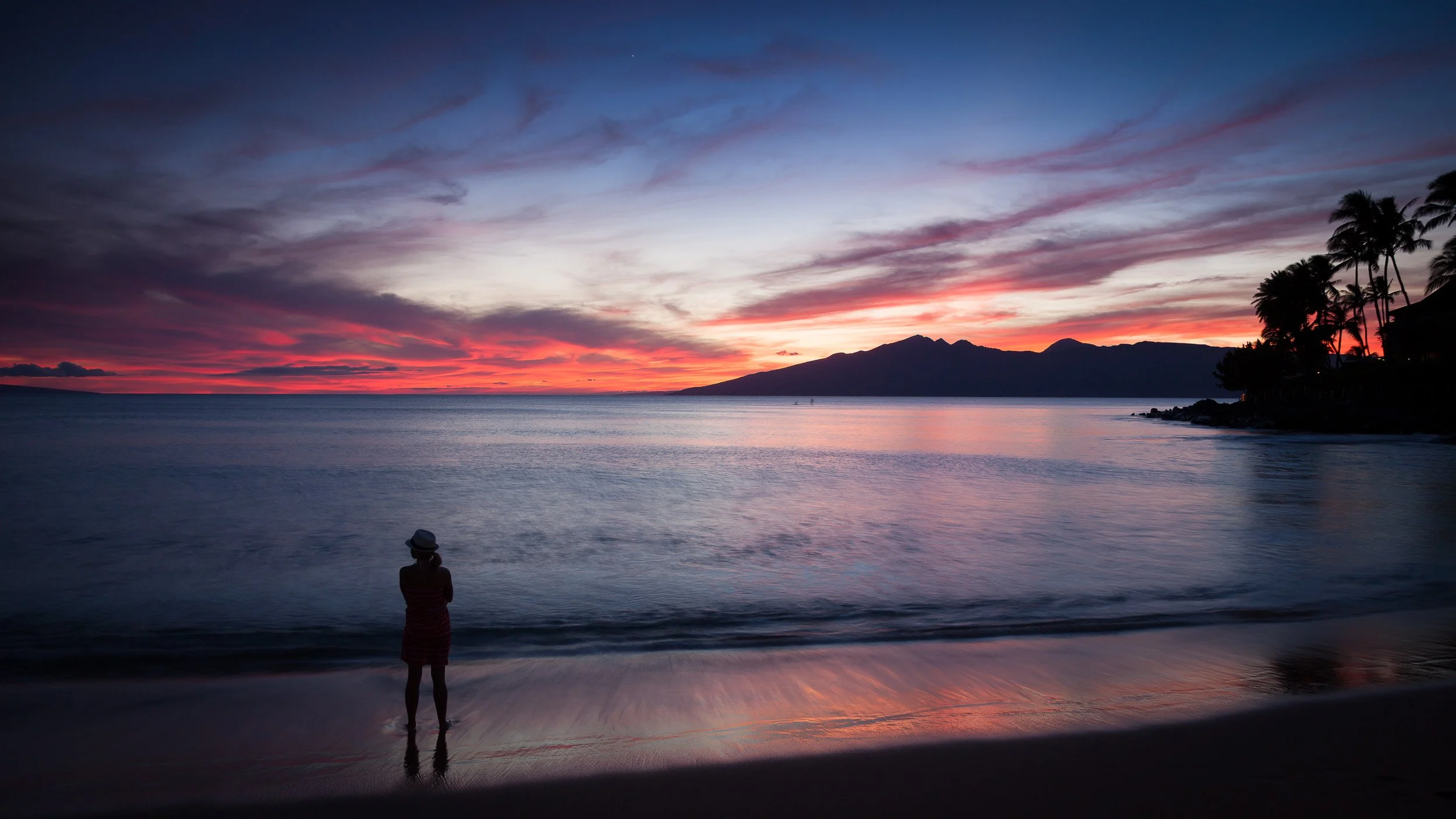 Sunset at Napili Kai on Maui, Hawaii, with a woman standing at the water’s edge as warm light spreads across the Pacific horizon.