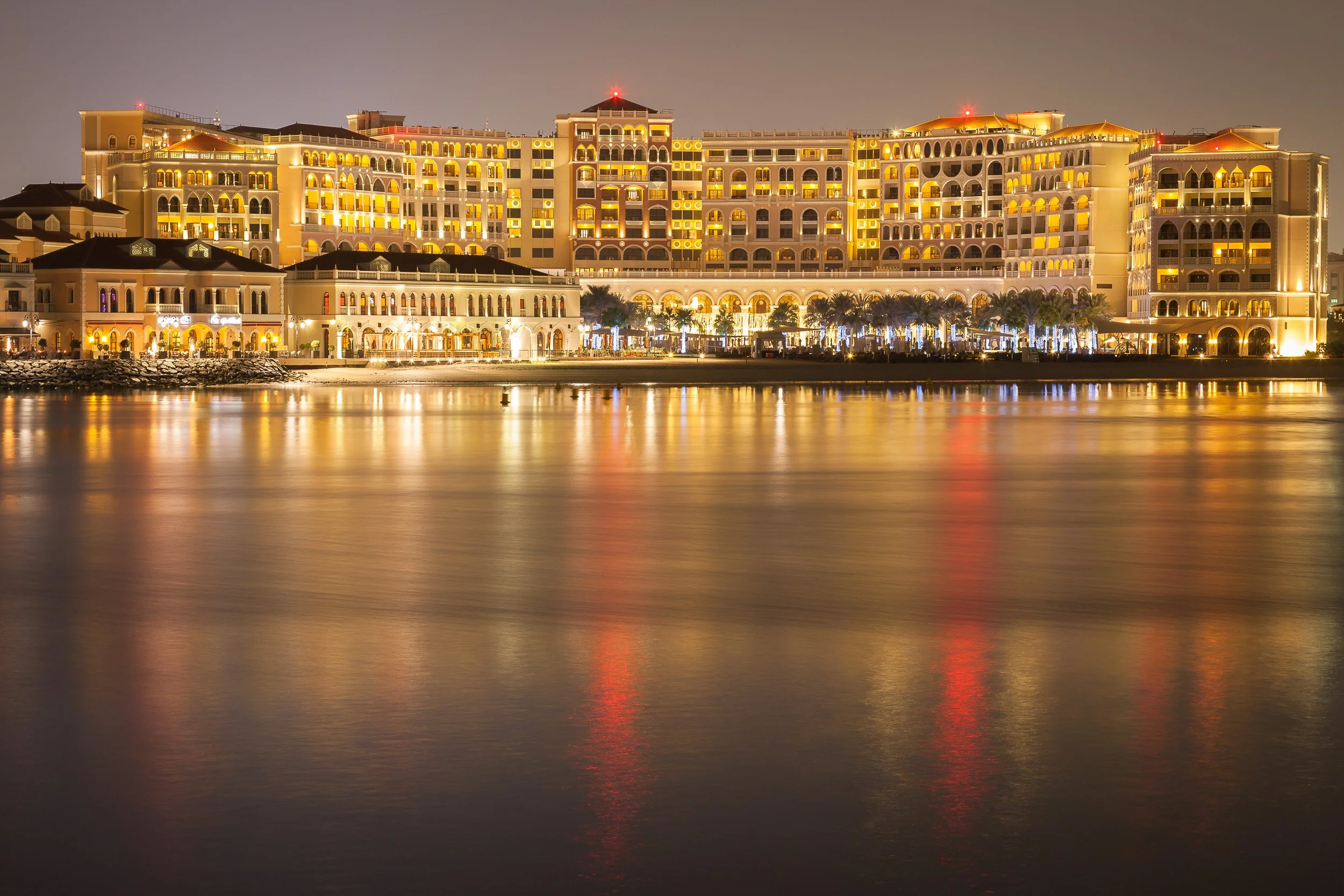 A grand waterfront hotel glowing softly across the water after nightfall, Abu Dhabi.