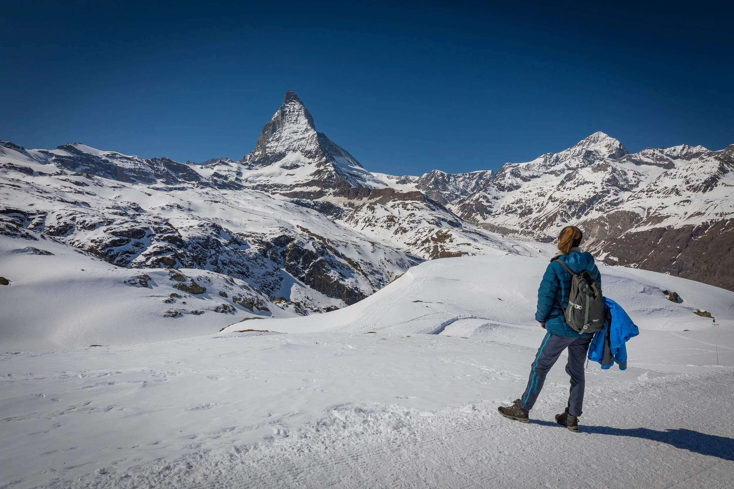 Blue-jacketed hiker pausing on a snowy Gornergrat ridge, looking out over the Matterhorn and a sea of jagged alpine peaks.
