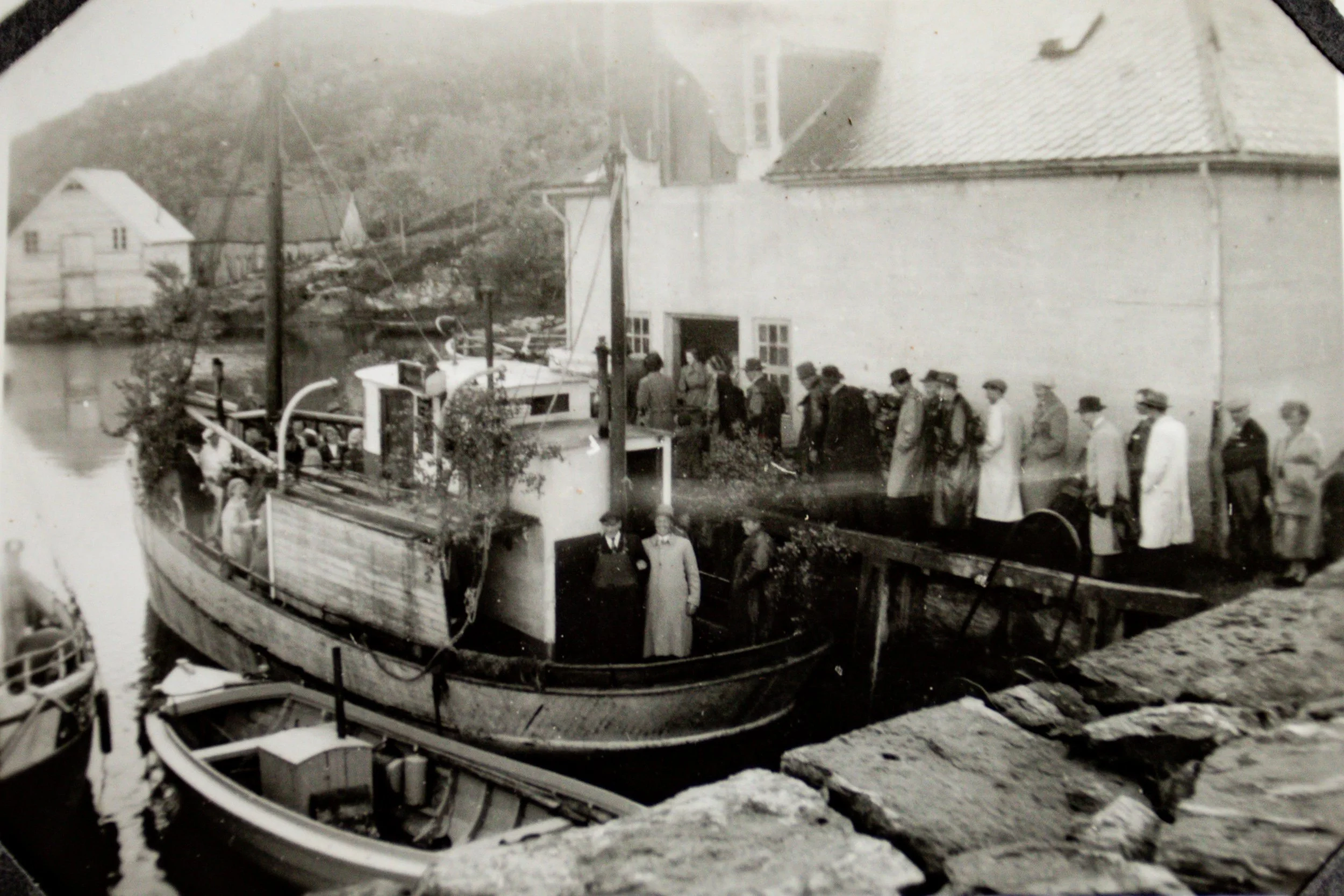«Til kirke 1952». Et bryllupsfølge samlet ved ishuset og moloen i Husevåg i forbindelse med reisen til kirken. En av kutterne fra Husevåg ligger ved kaia, pyntet med løvkvister langs overbygningen, mens gjestene står samlet både på land og om bord. H